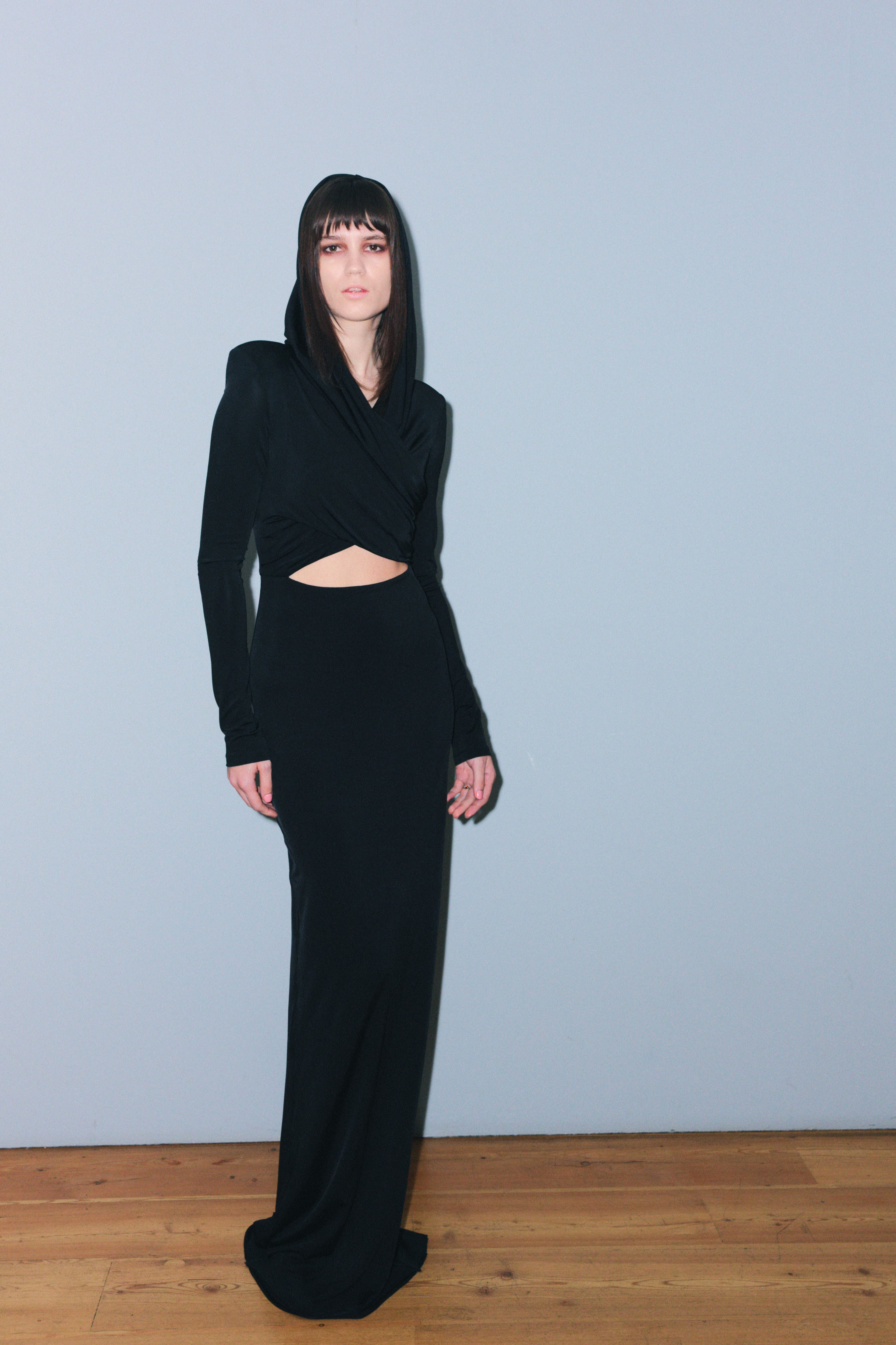 Элегантное черное платье с капюшоном и вырезом на талии, подчеркивающее силуэт, создает атмосферу загадочности и стиля.