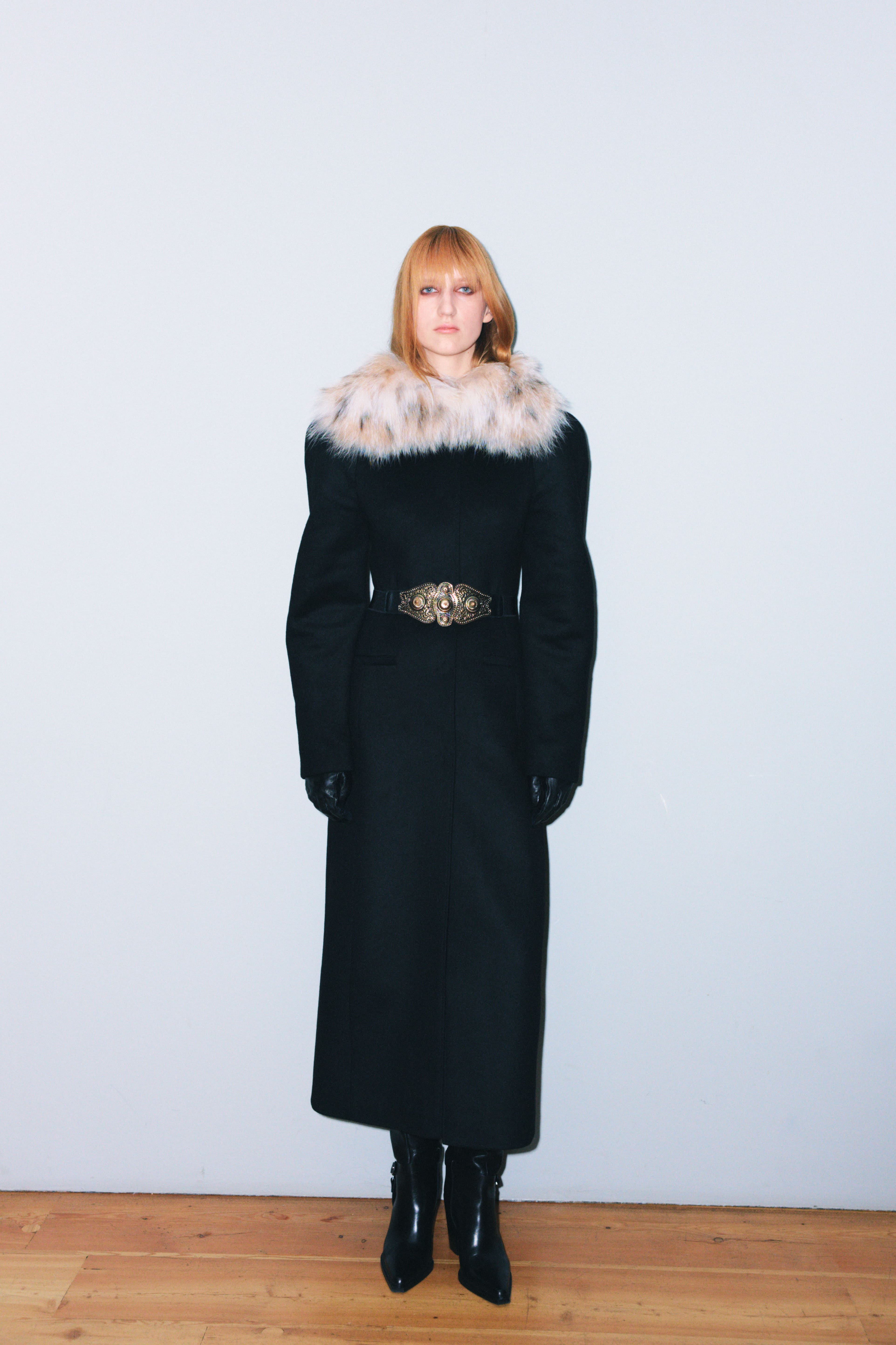 Элегантное черное пальто с меховым воротником и золотым поясом, создающее контраст с гладкими кожаными перчатками и ботфортами.