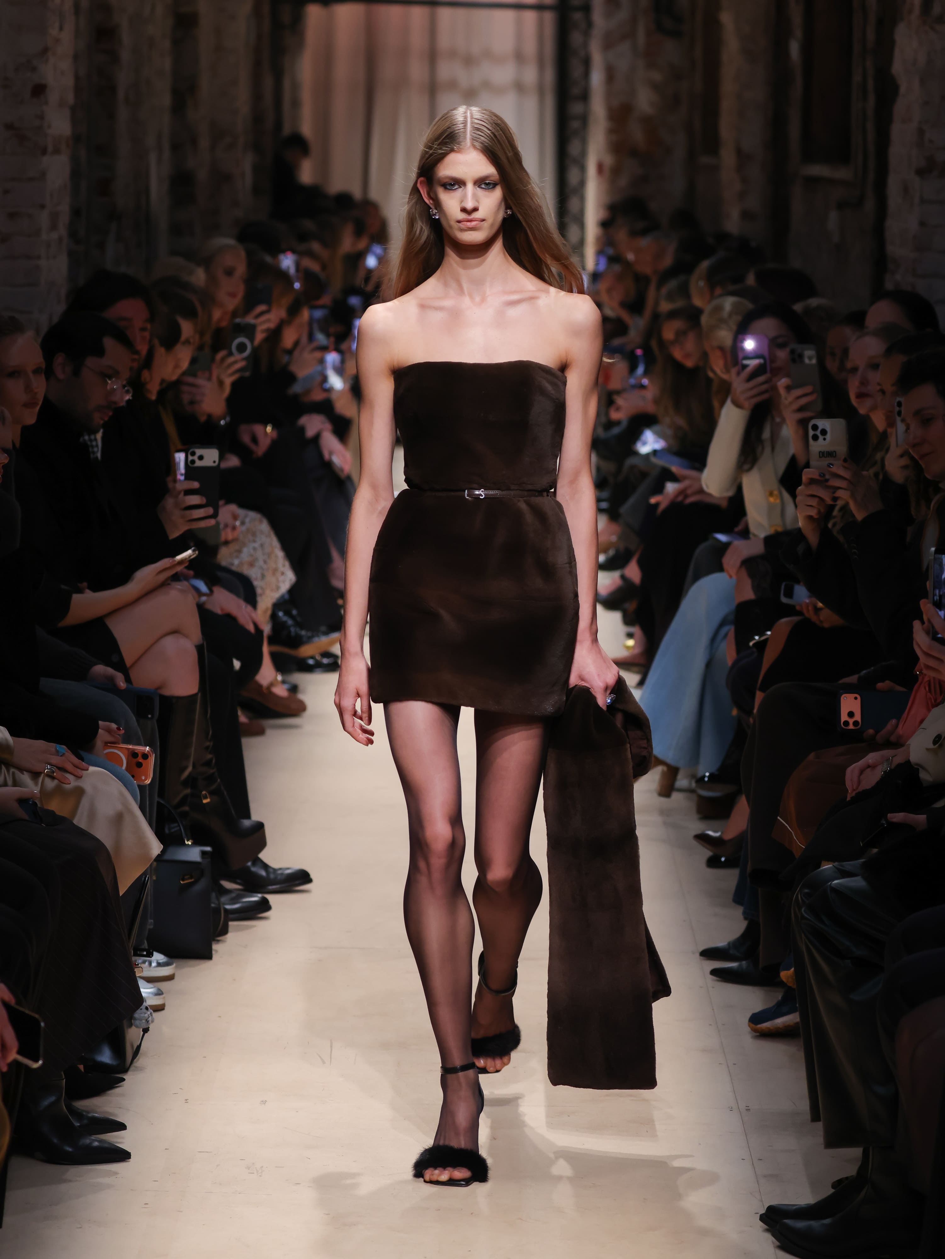 Модель в облегающем коричневом мини-платье с прямыми линиями и мягкой текстурой, дополняет образ меховым аксессуаром.