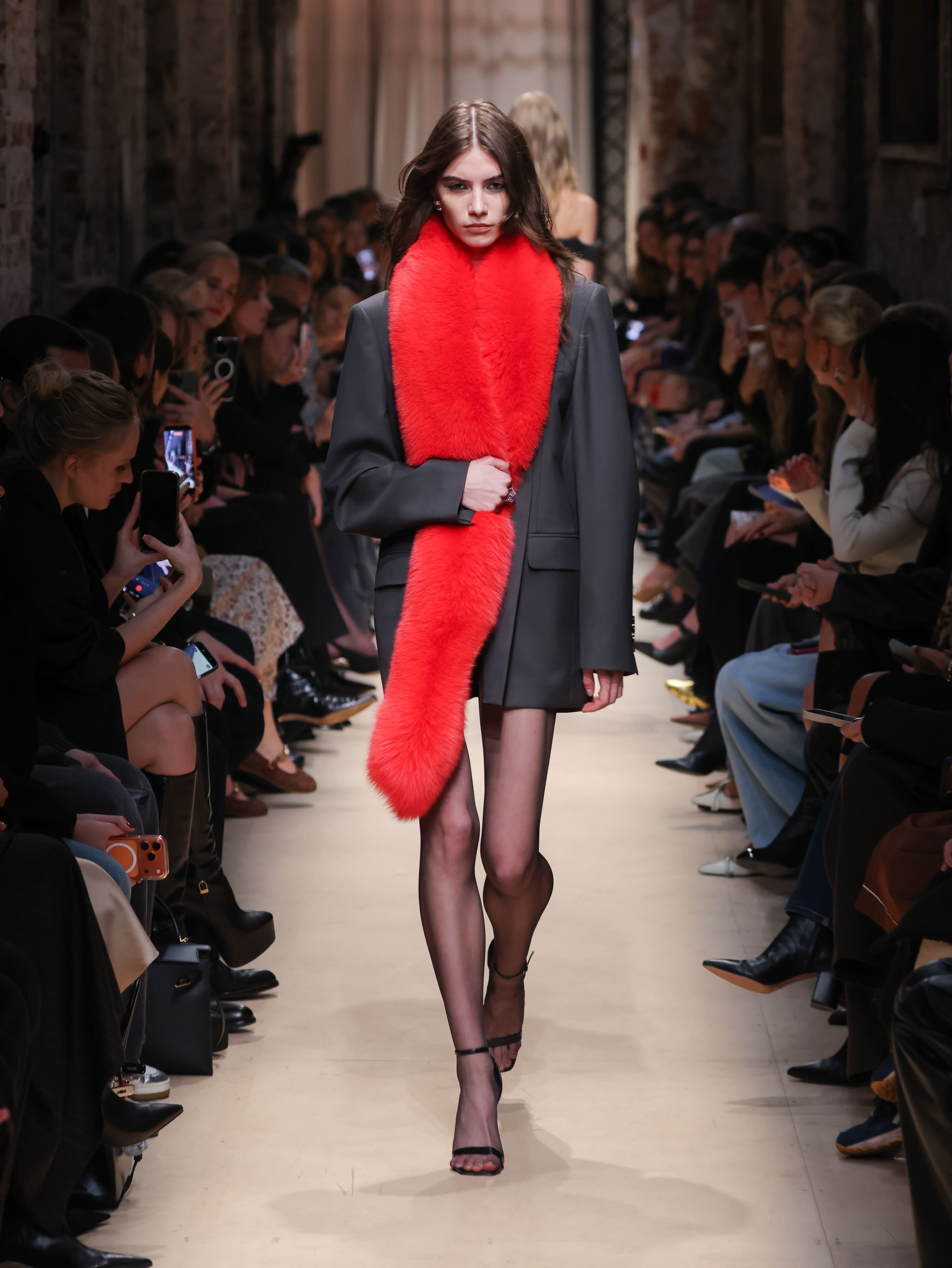 Модель в строгом сером жакете и мини-юбке, обрамленная ярким красным меховым шарфом, уверенно шагает по подиуму.