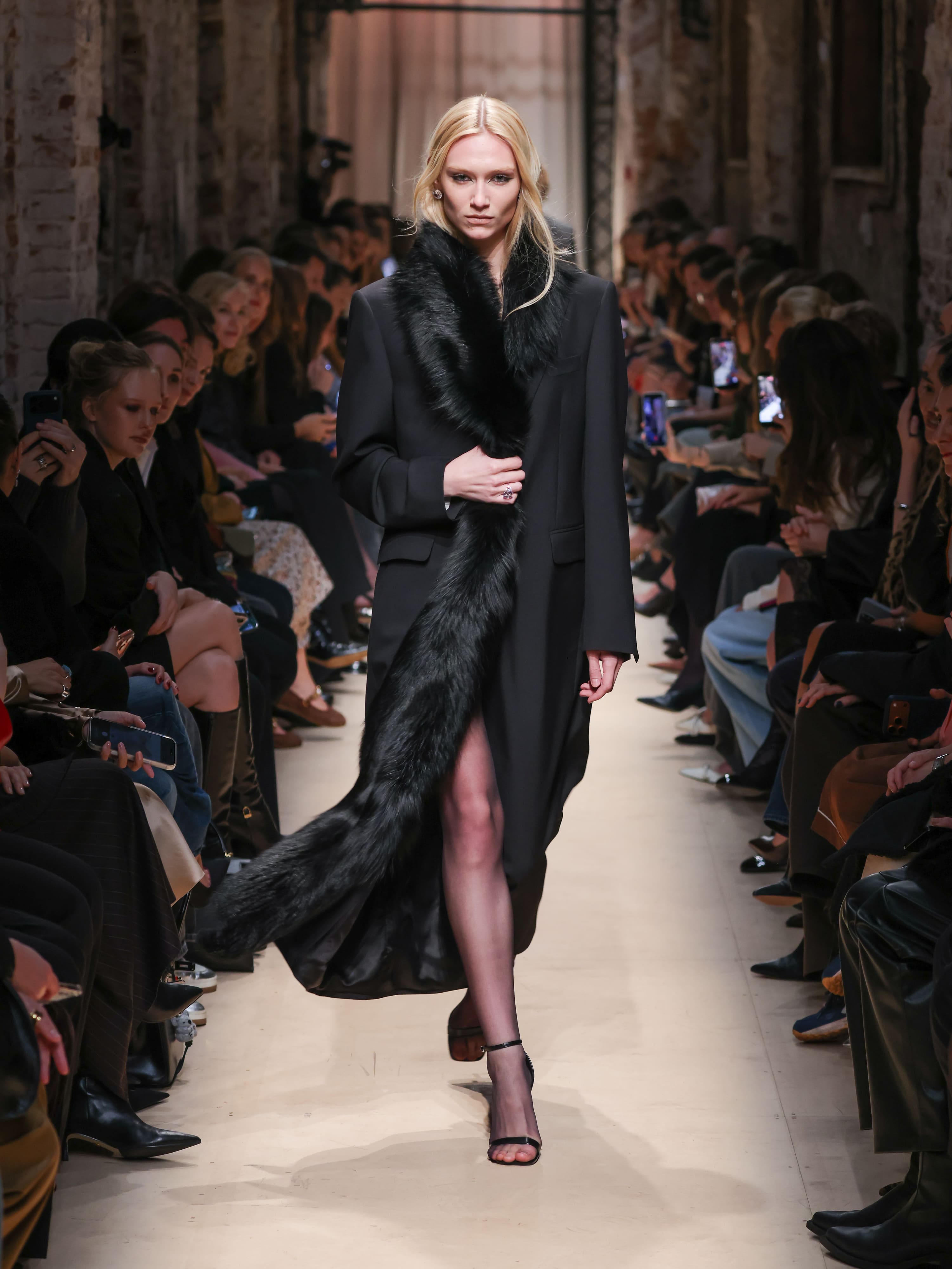 Элегантное черное пальто с длинным меховым шарфом, подчеркивающее стройную фигуру модели, создает атмосферу утонченной роскоши.