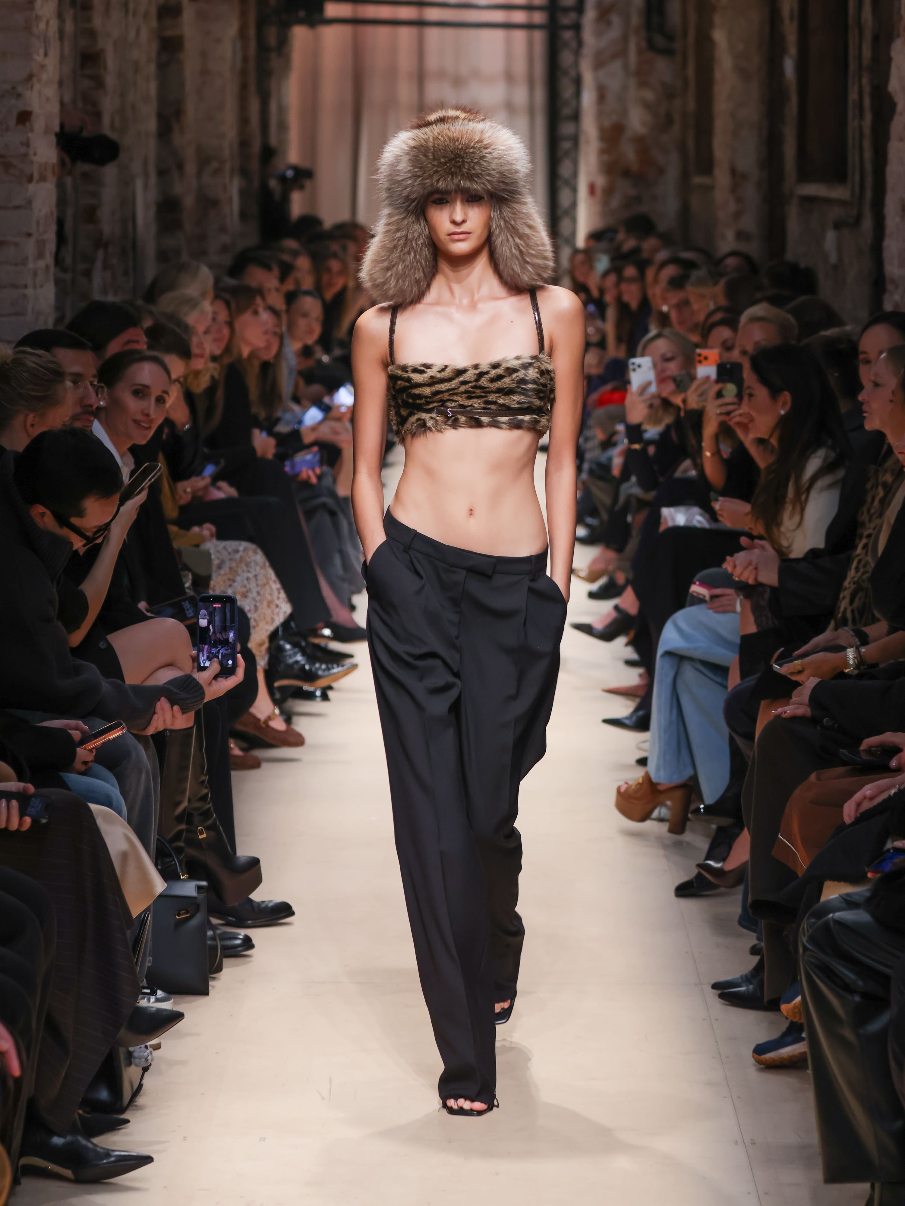 Модель в леопардовом топе и черных широких брюках, завершает образ меховой шапкой, создавая эффектный контраст на подиуме.