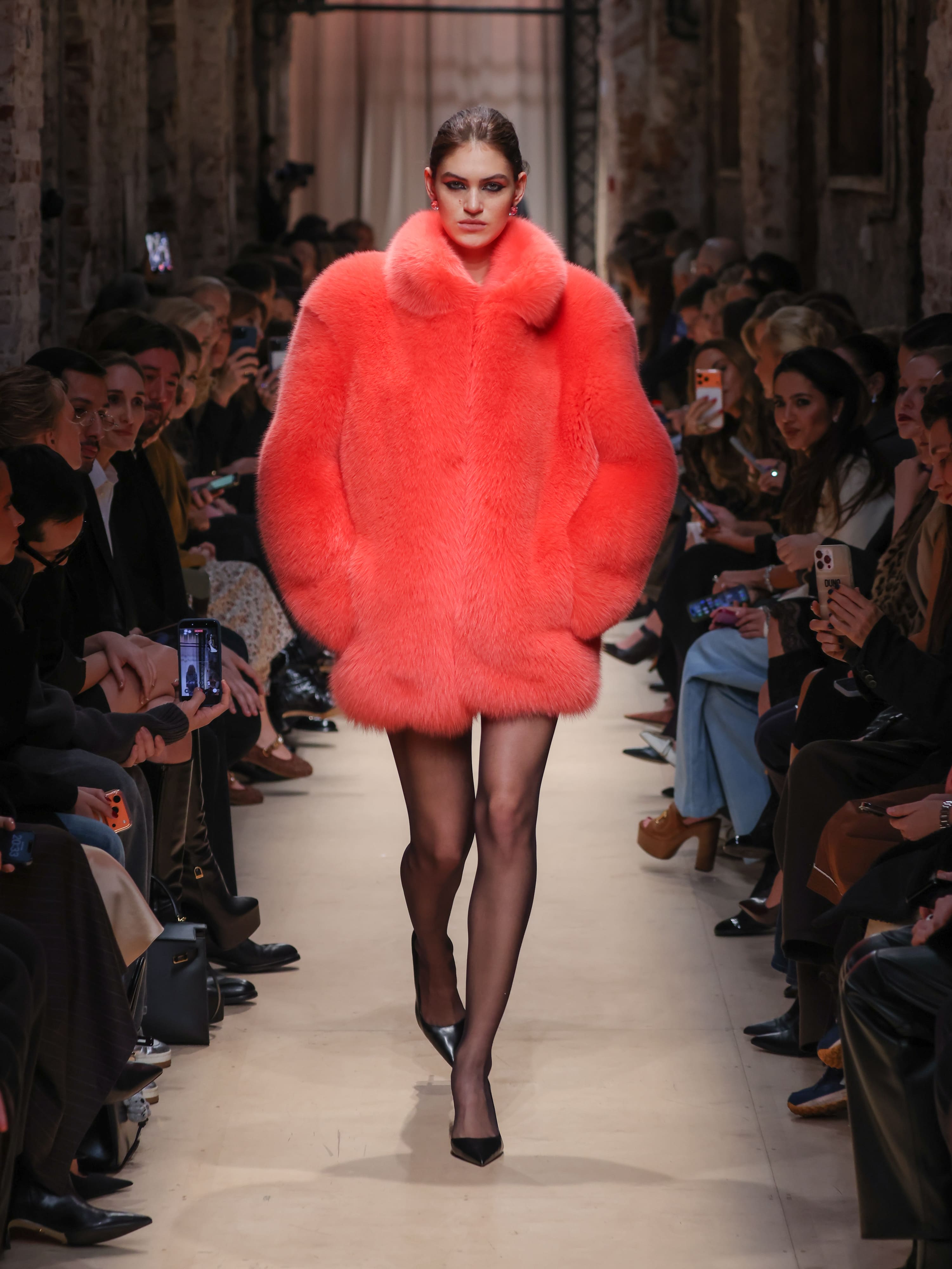 Модель в ярком коралловом пуховике с объемным воротником и блестящими чулками, уверенно шагает по подиуму среди зрителей.