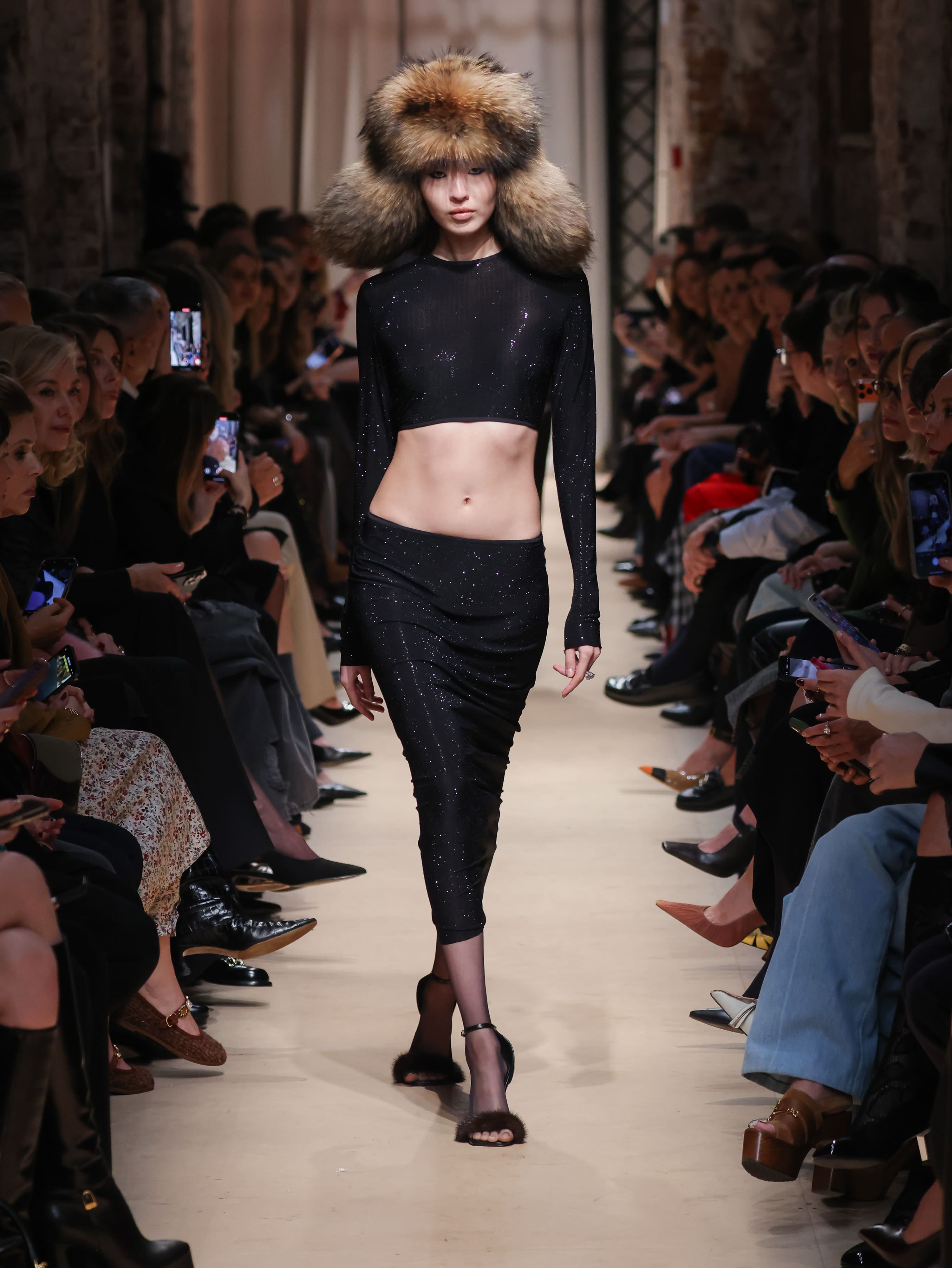 Модель в черном наряде с блестками и обтягивающей юбкой, дополненная объемной шапкой из меха, уверенно шагает по подиуму.