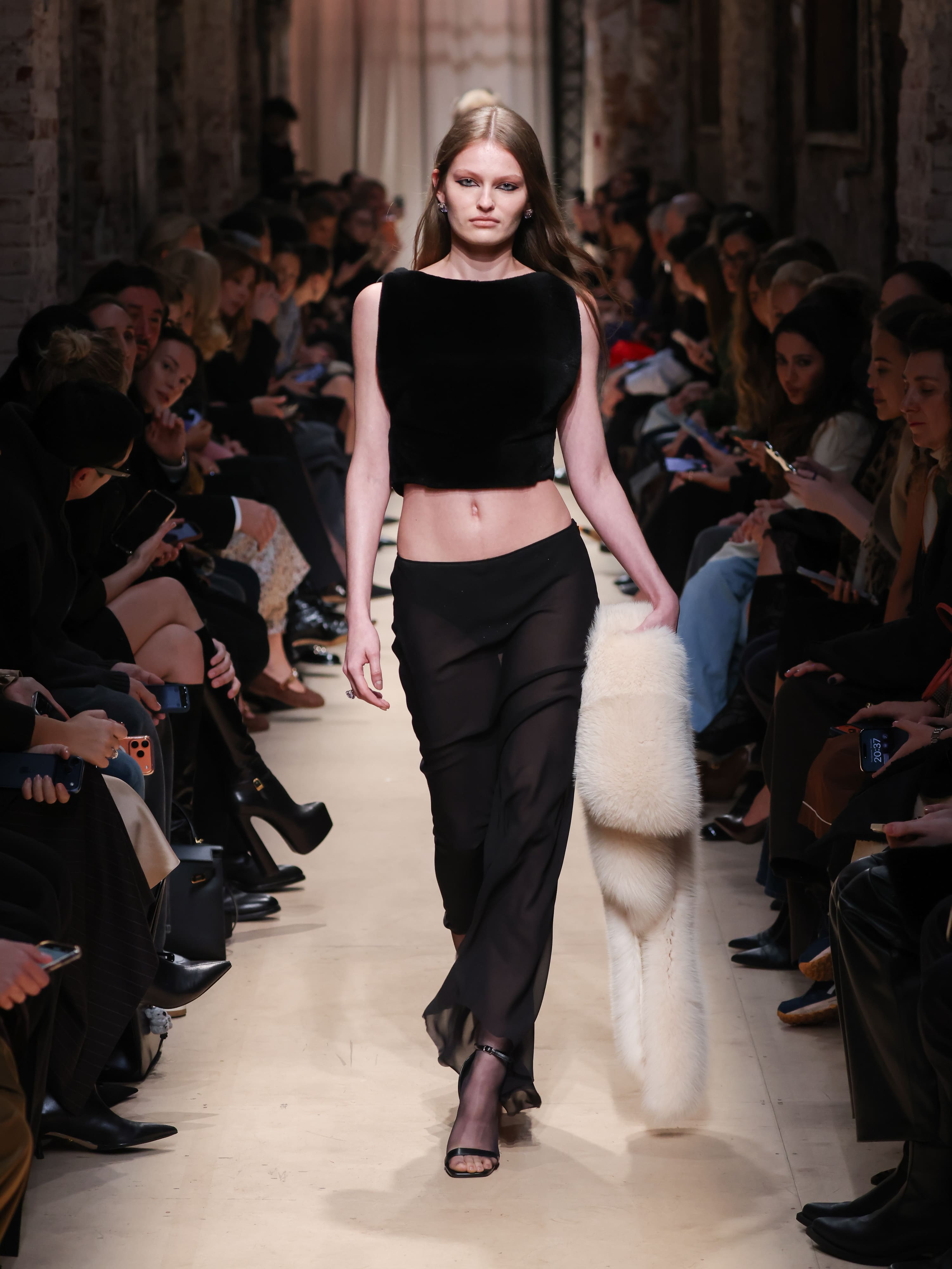 Модель в черном бархатном топе и полупрозрачных брюках, с белым меховым палантином, уверенно шагает по подиуму осени/зимы 2026 от Sashaverse