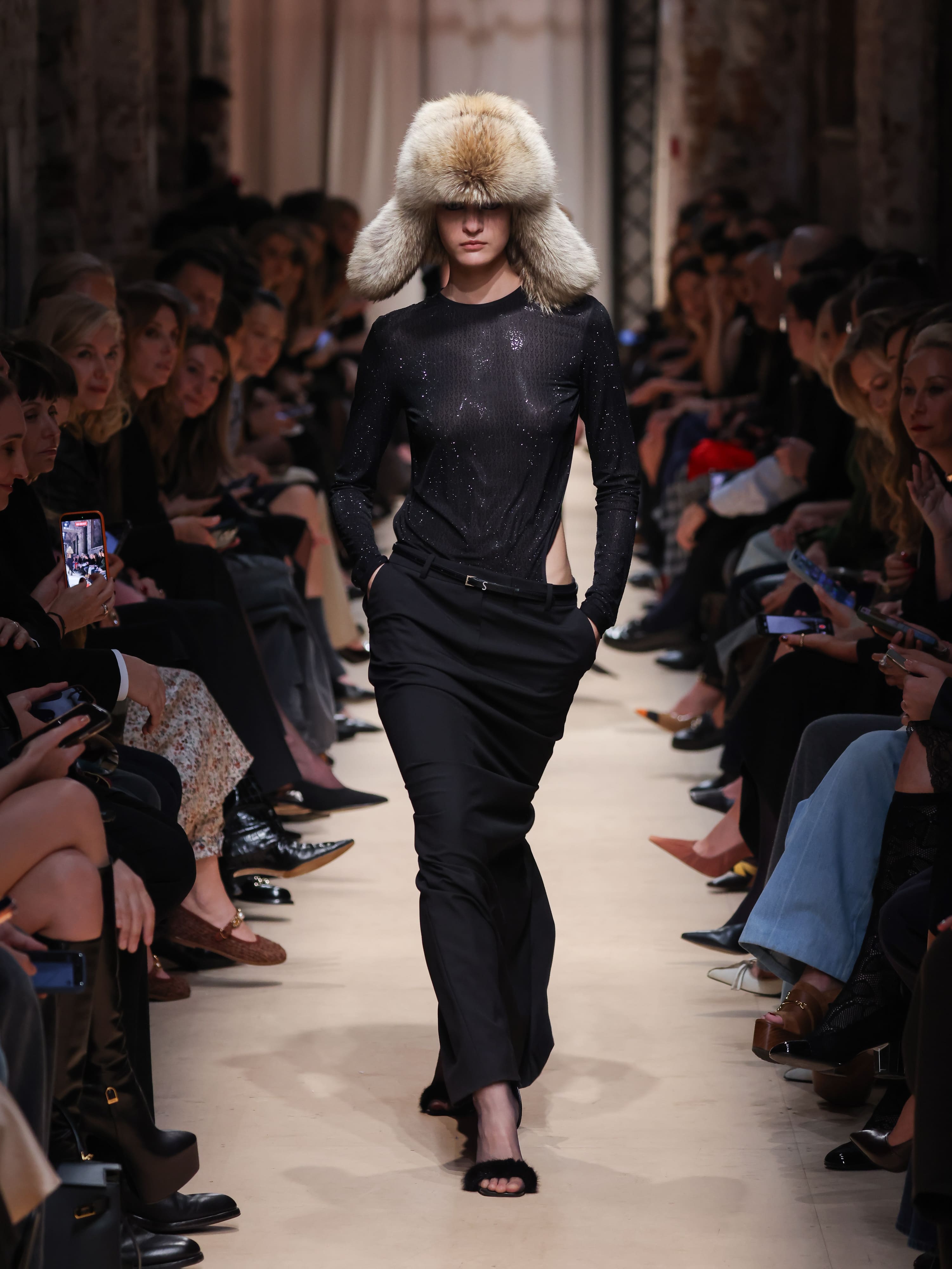 Модель в черной облегающей блузе с блестками и широких брюках, дополненная объемной меховой шапкой, создает атмосферу зимней элегантности....