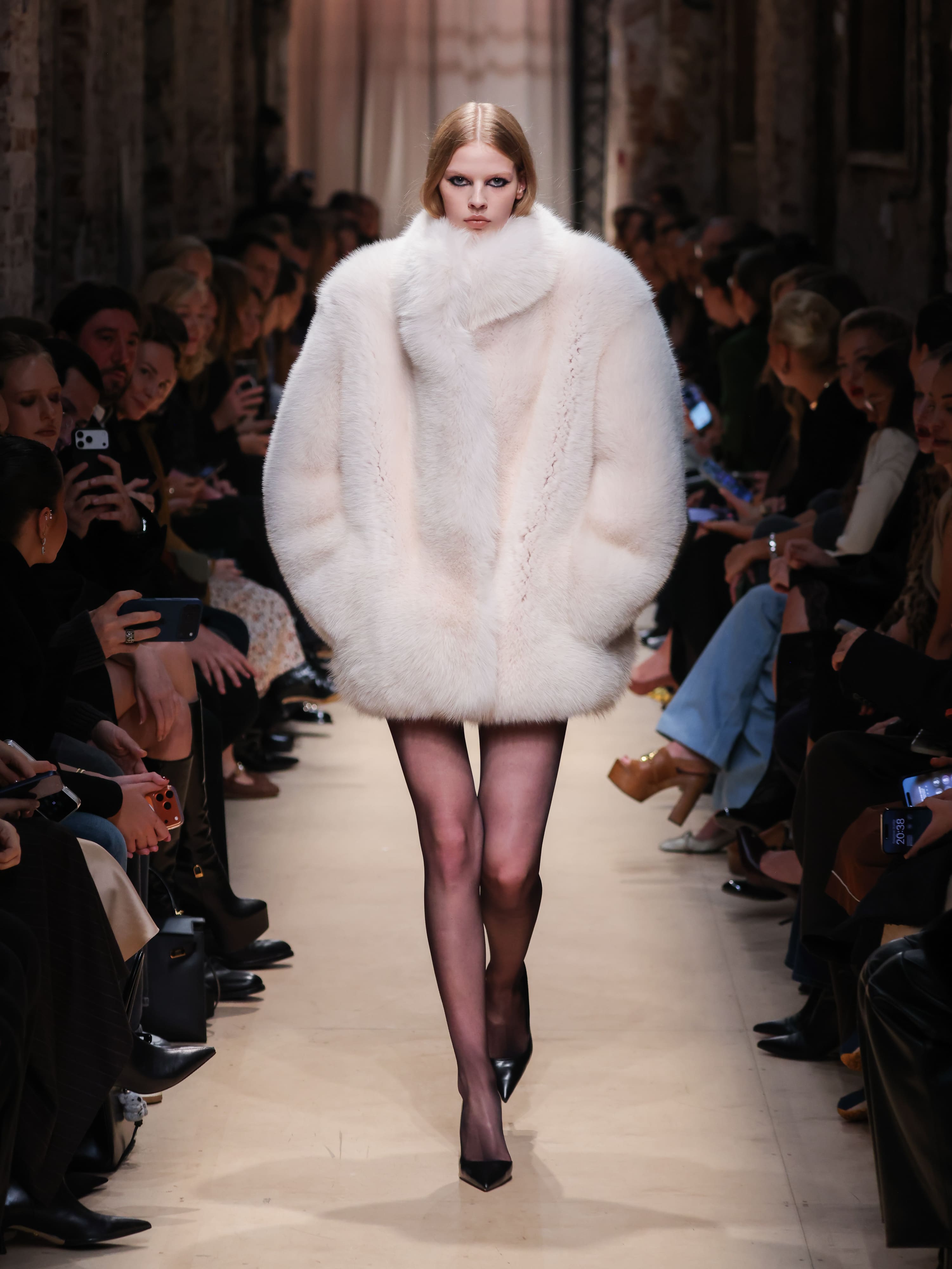 Модель в объемном светлом меховом пальто с высоким воротником, сочетая элегантность и уют, уверенно шагает по подиуму.