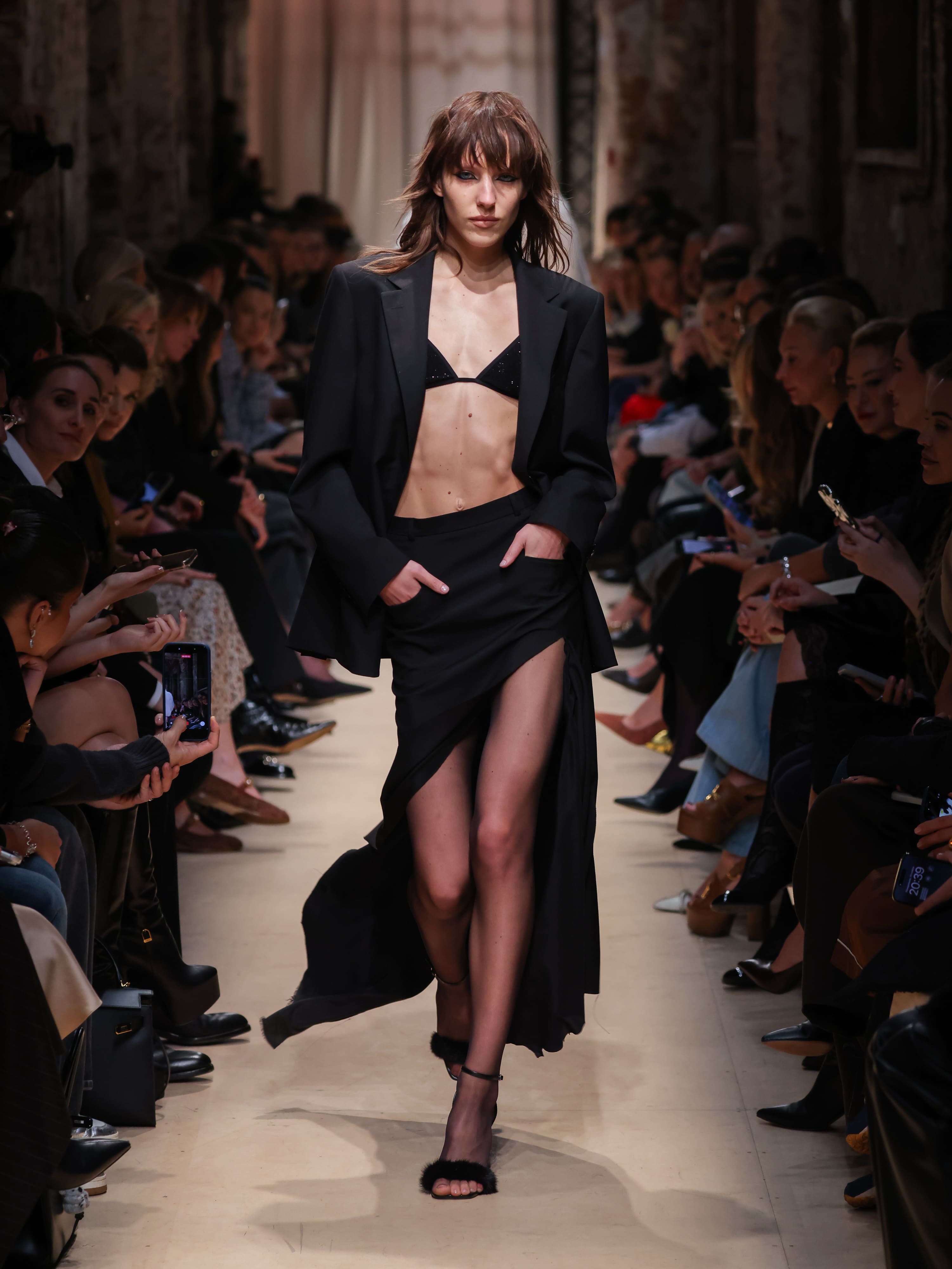 Модель в черном костюме с открытым топом и высокой юбкой, подчеркивающей фигуру, уверенно шагает по подиуму осени-зимы 2026 от Sashaverse.