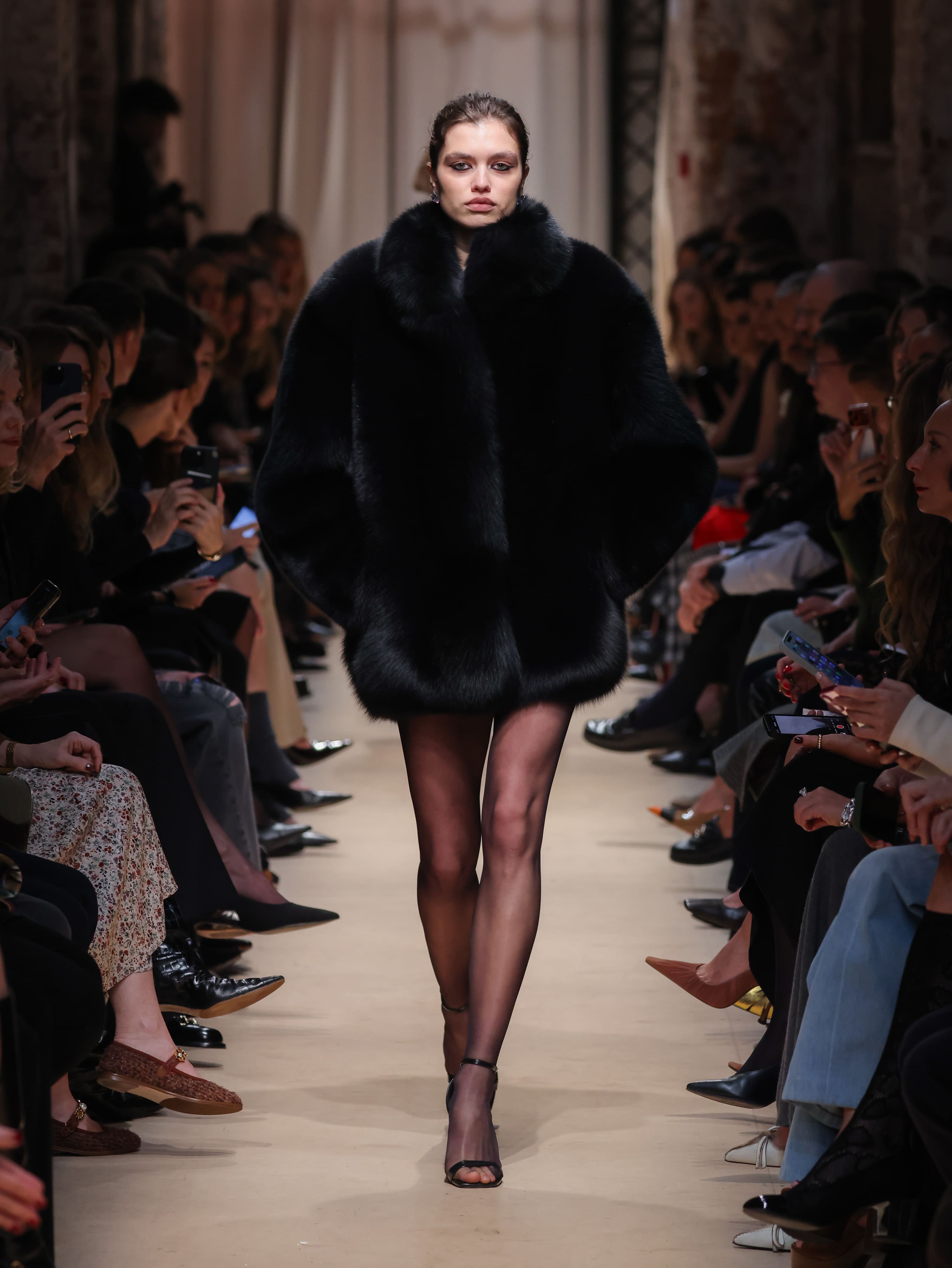 Модель в объемном черном меховом пальто с высоким воротником, сочетая элегантность и смелость, уверенно шагает по подиуму.