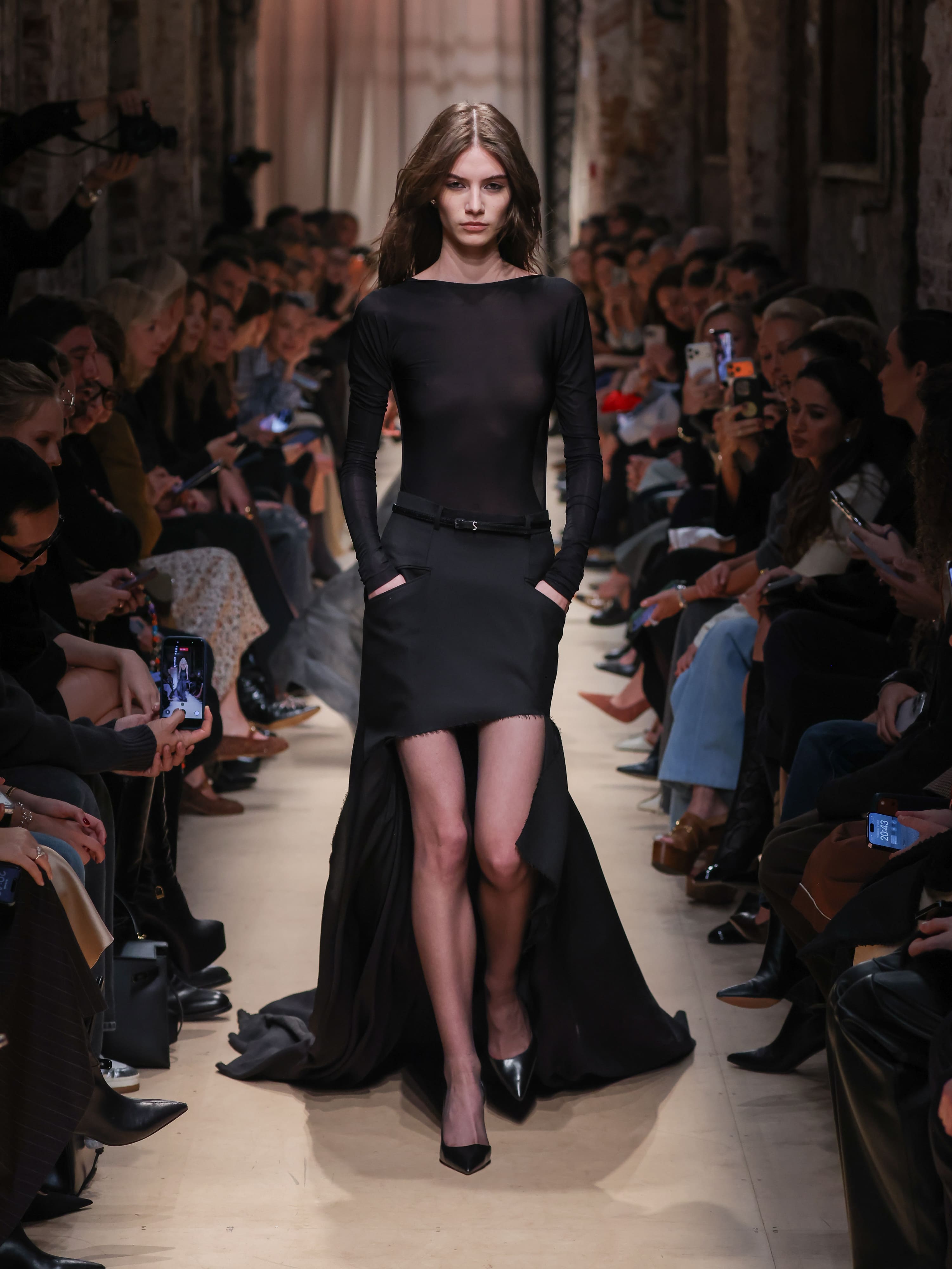 Модель в полупрозрачном черном топе и асимметричной юбке с высоким разрезом, элегантно шагает по подиуму, создавая атмосферу загадочности....