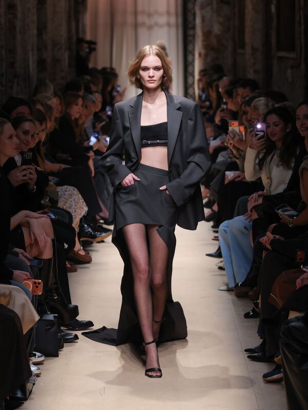 Элегантный образ от Sashaverse: черный жакет с открытым топом и асимметричной юбкой, подчеркивающий силуэт модели на фоне зрителей.