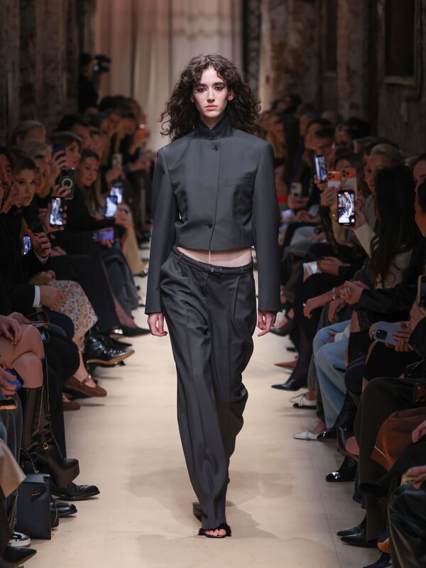 Модель в строгом сером костюме с укороченным жакетом и широкими брюками, уверенно шагает по подиуму среди зрителей.