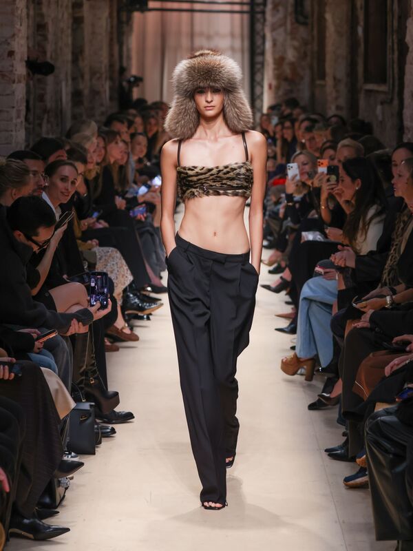 Модель в леопардовом топе и черных широких брюках, завершает образ меховой шапкой, создавая эффектный контраст на подиуме.