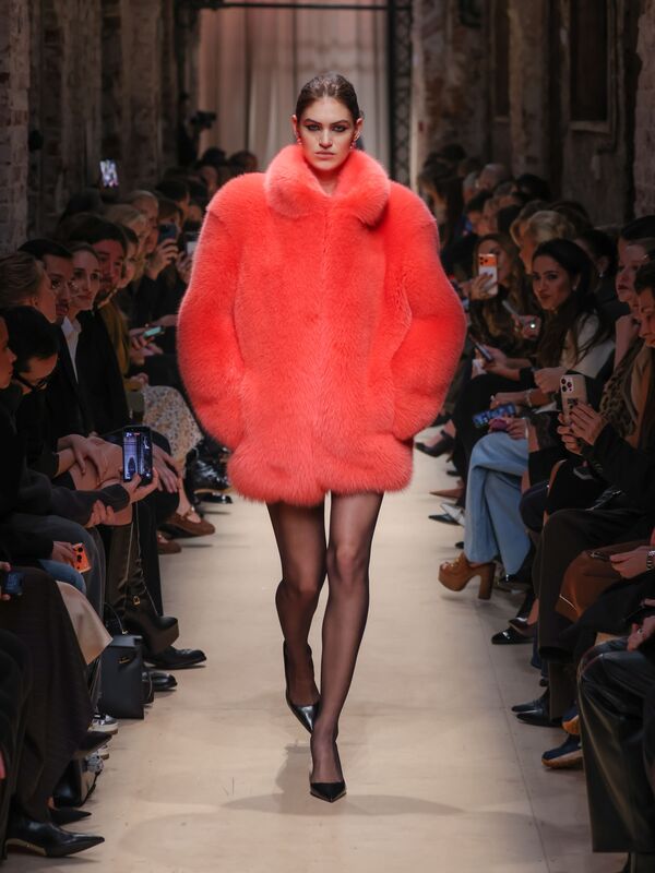 Модель в ярком коралловом пуховике с объемным воротником и блестящими чулками, уверенно шагает по подиуму среди зрителей.