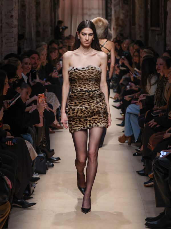 Модель в облегающем платье с леопардовым принтом и меховой отделкой, уверенно шагает по подиуму, привлекая взгляды зрителей.