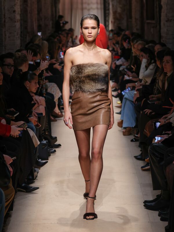 Модель в коротком кожаном платье с меховым верхом, уверенно шагает по подиуму, создавая атмосферу элегантной дерзости.