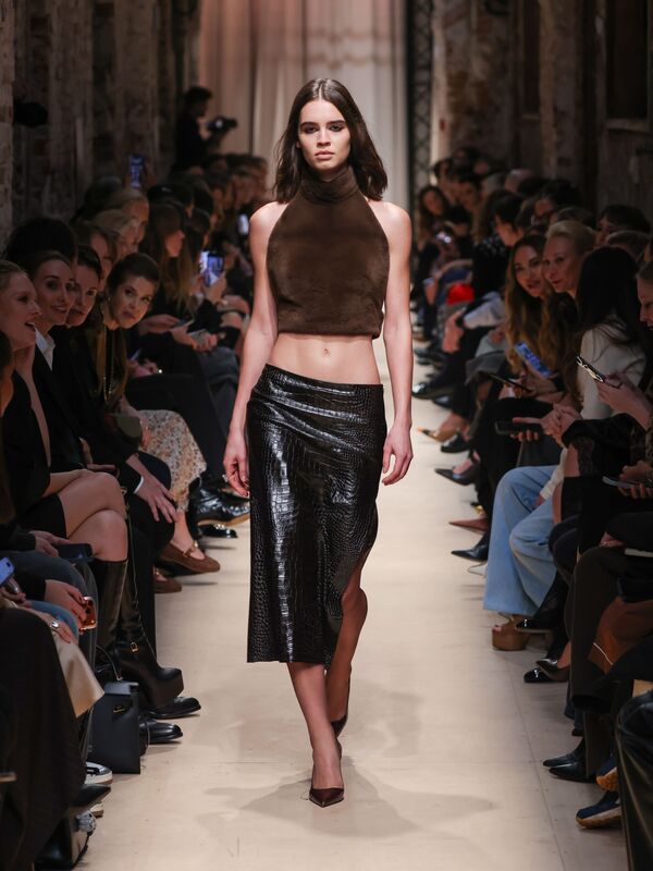 Модель в коричневом топе и блестящей юбке с текстурой крокодила уверенно шагает по подиуму, привлекая взгляды зрителей.