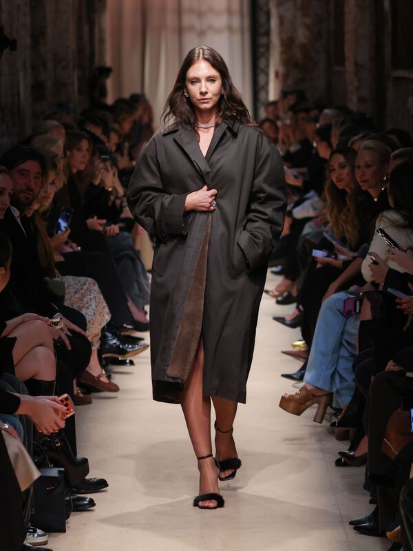 Модель в объемном темно-коричневом пальто с контрастной отделкой, уверенно шагает по подиуму, создавая атмосферу утонченной элегантности.
