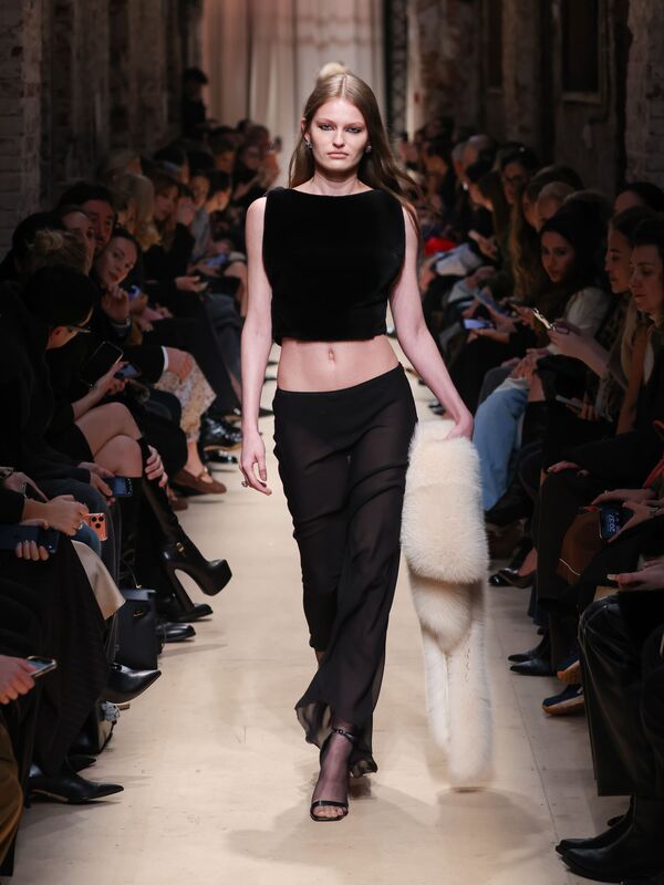 Модель в черном бархатном топе и полупрозрачных брюках, с белым меховым палантином, уверенно шагает по подиуму осени/зимы 2026 от Sashaverse