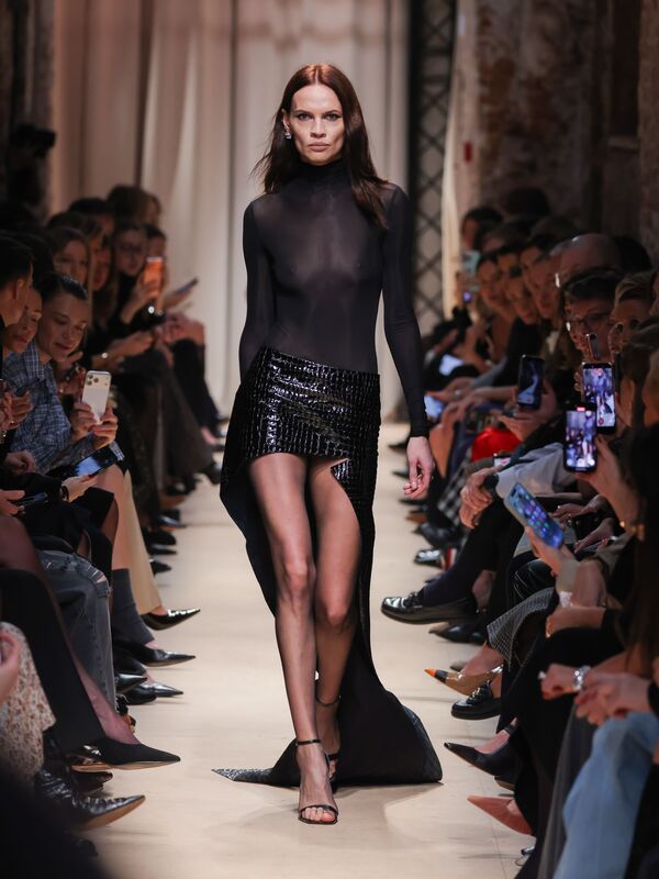 Модель в полупрозрачном черном топе и блестящей юбке с высоким разрезом демонстрирует элегантность и смелость коллекции Sashaverse осень-зима 2026....