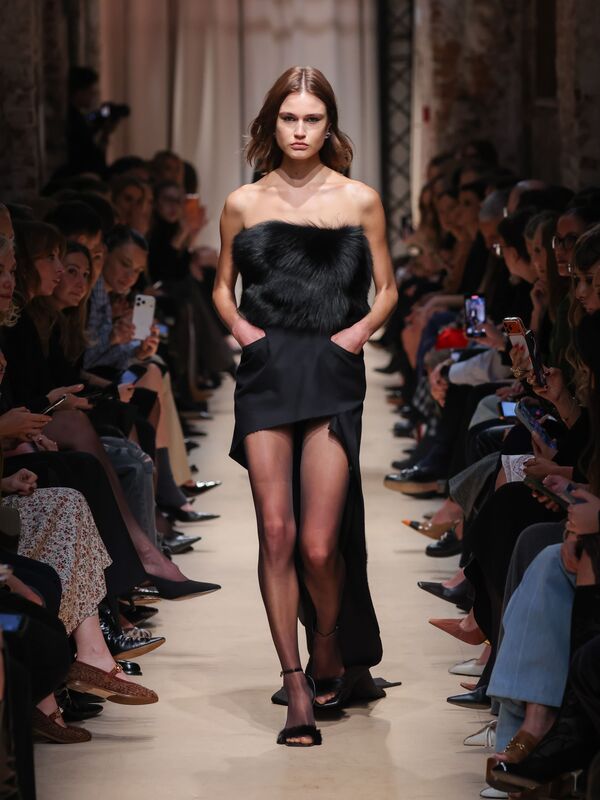 Элегантное черное платье с объемным меховым верхом и разрезом, подчеркивающее стройные ноги модели, создает атмосферу утонченной роскоши.