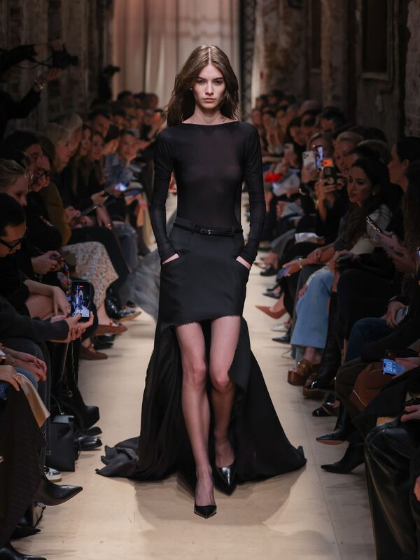 Модель в полупрозрачном черном топе и асимметричной юбке с высоким разрезом, элегантно шагает по подиуму, создавая атмосферу загадочности....