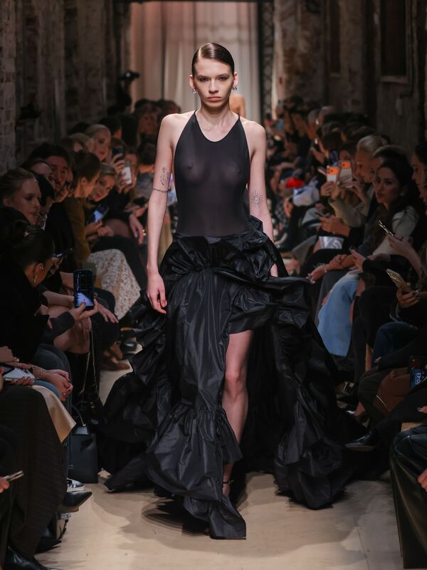 Модель в полупрозрачном черном наряде с объемной юбкой из текстурированной ткани, создающей эффект драмы на подиуме.
