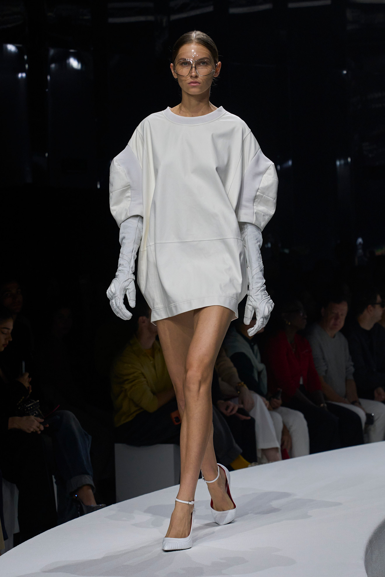 Модель в объемном белом платье с асимметричными рукавами и длинных перчатках, завершает образ сандалиями на низком каблуке.