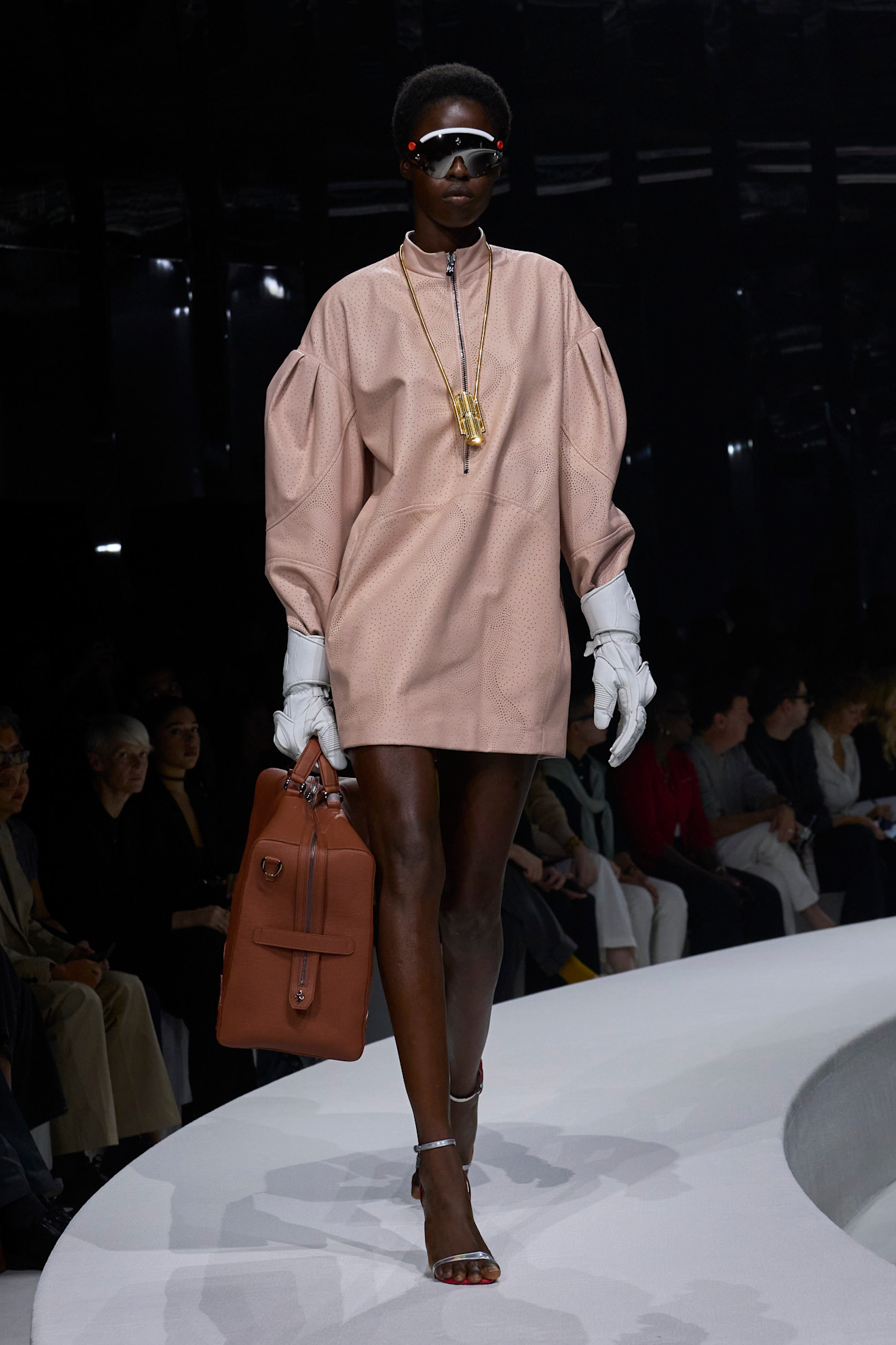 Модель в розовом платье с объемными рукавами, дополненном белыми перчатками и стильными очками, несет коричневую сумку.