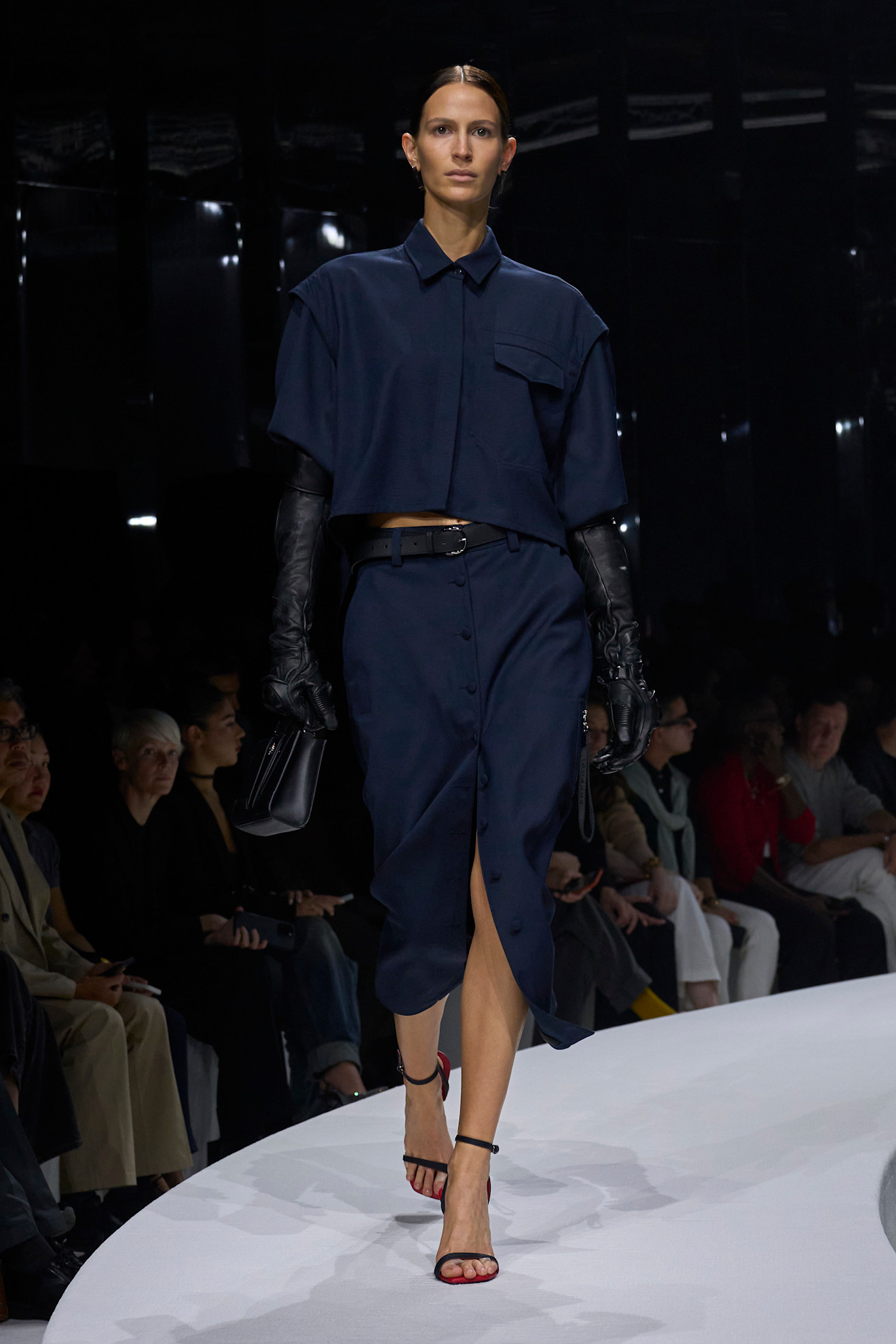 Модель в темно-синем платье-рубашке с короткими рукавами и разрезом, дополненная кожаными перчатками и элегантными босоножками на тонком каблуке....