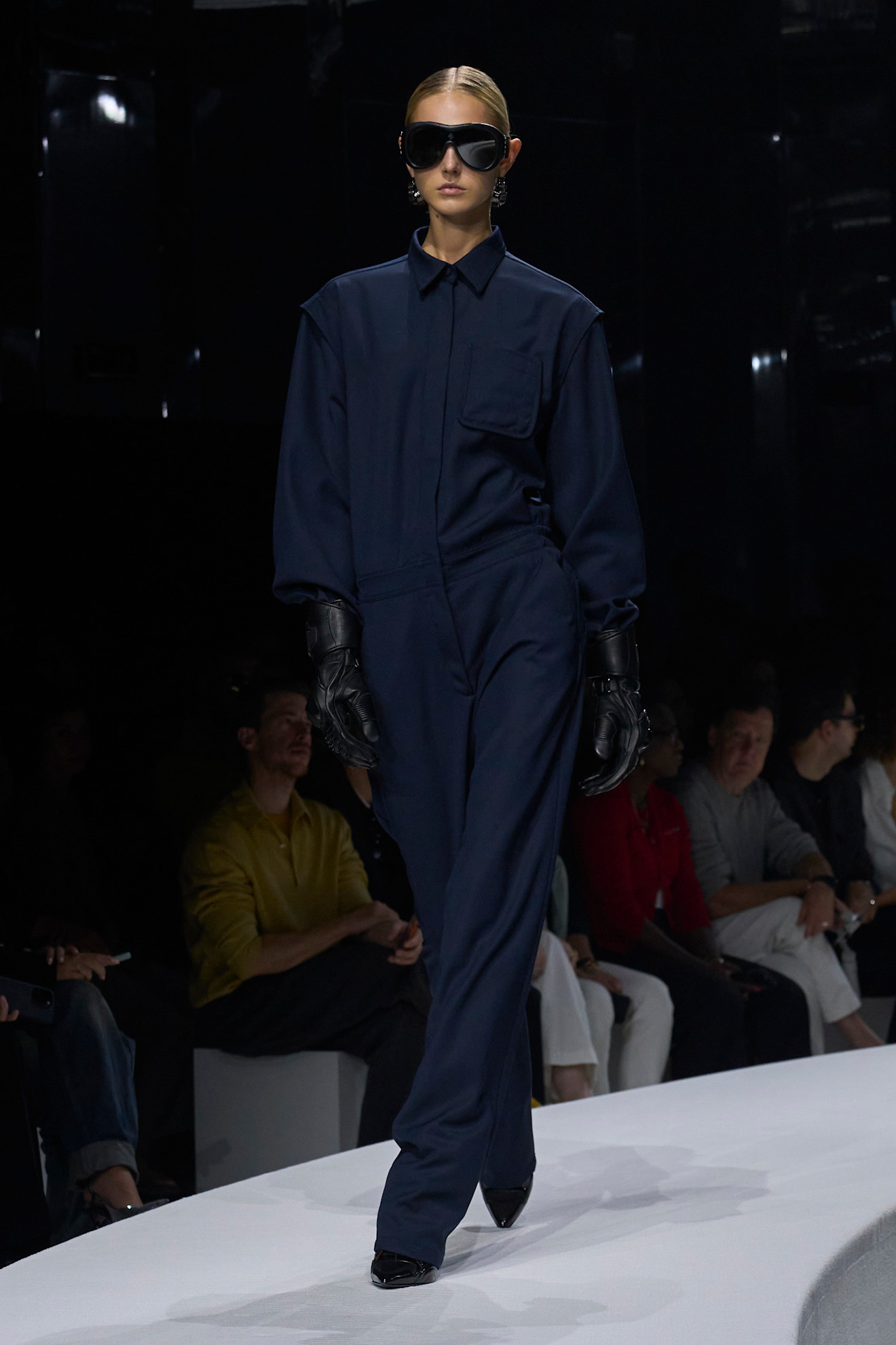 Модель в темно-синем комбинезоне с объемными рукавами и кожаными перчатками, завершает образ стильными солнцезащитными очками.