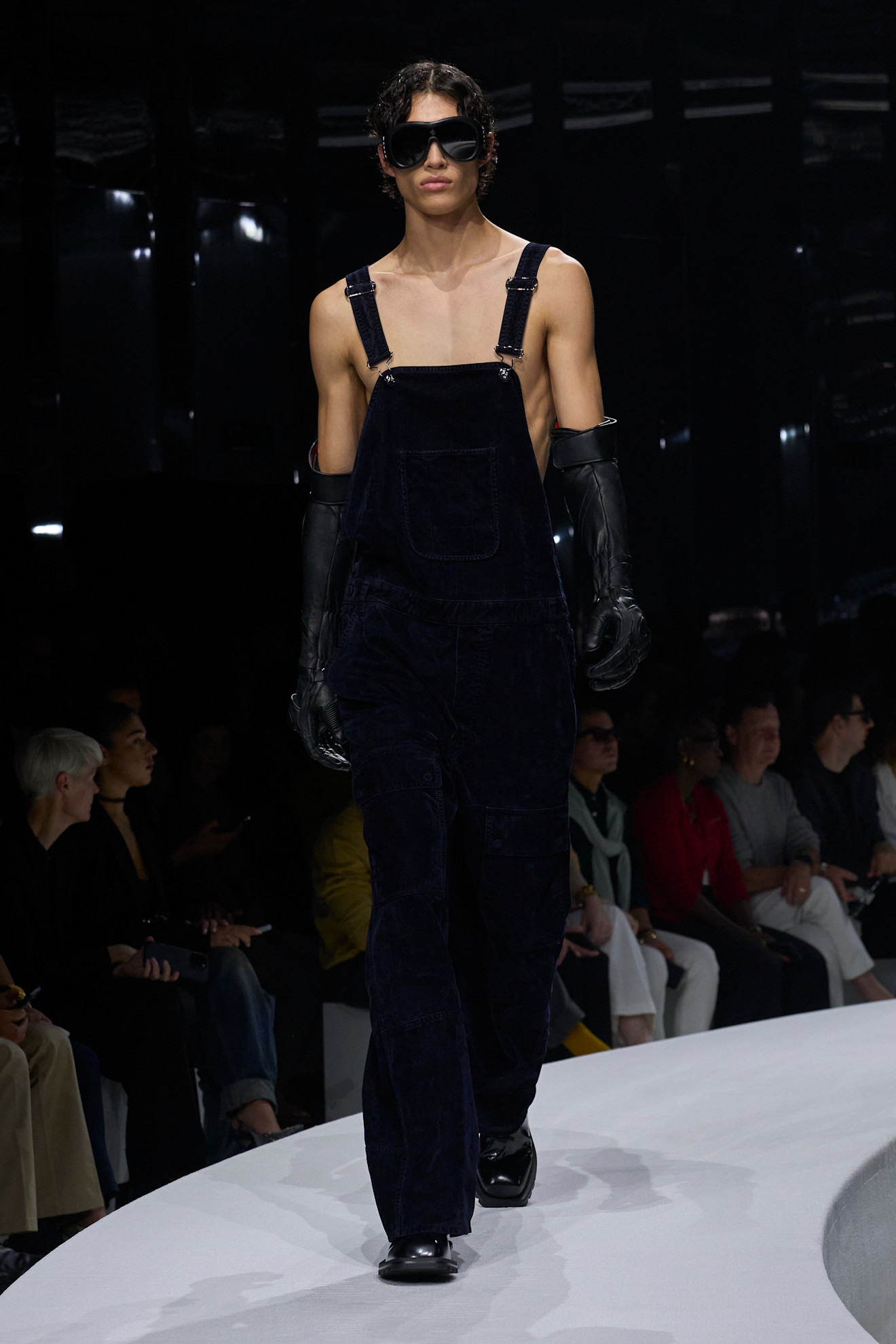 Модель в темно-синих вельветовых комбинезонах с открытыми плечами и кожаными перчатками, создающих эффект дерзкого минимализма.