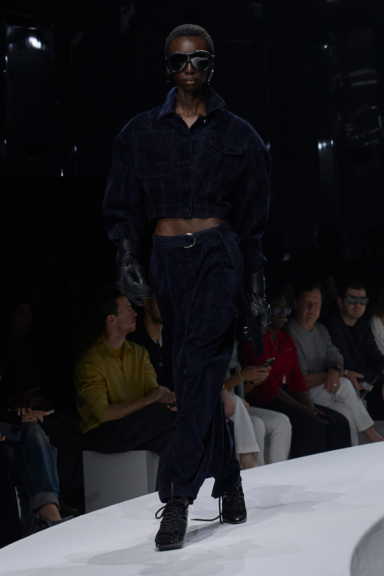 Модель в темно-синем велюровом комплекте с укороченной курткой и широкими брюками, дополненная массивными очками и перчатками.