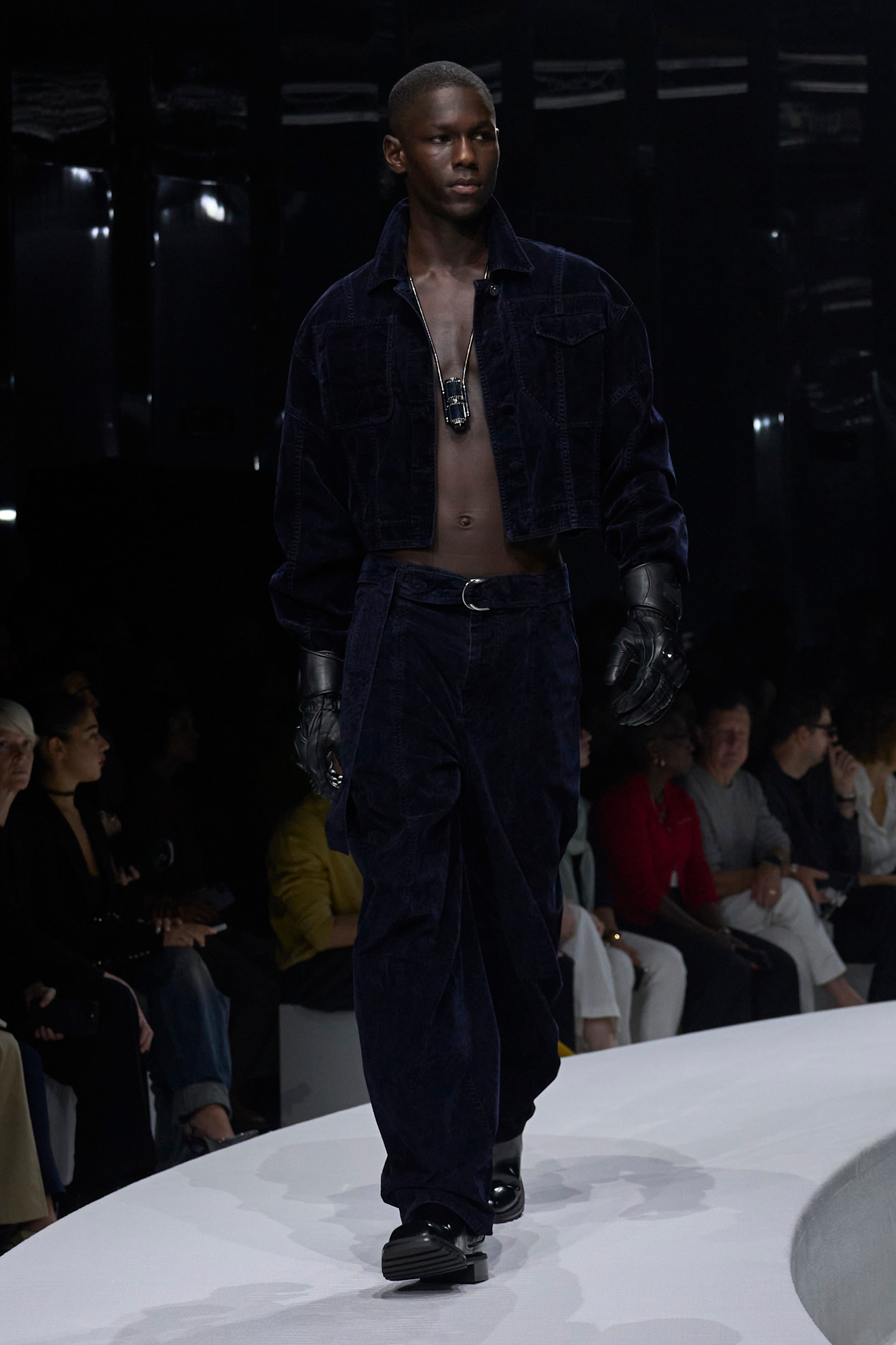 Модель в темно-синем велюровом костюме с укороченной курткой и широкими брюками, дополненная кожаными перчатками и массивной цепью.