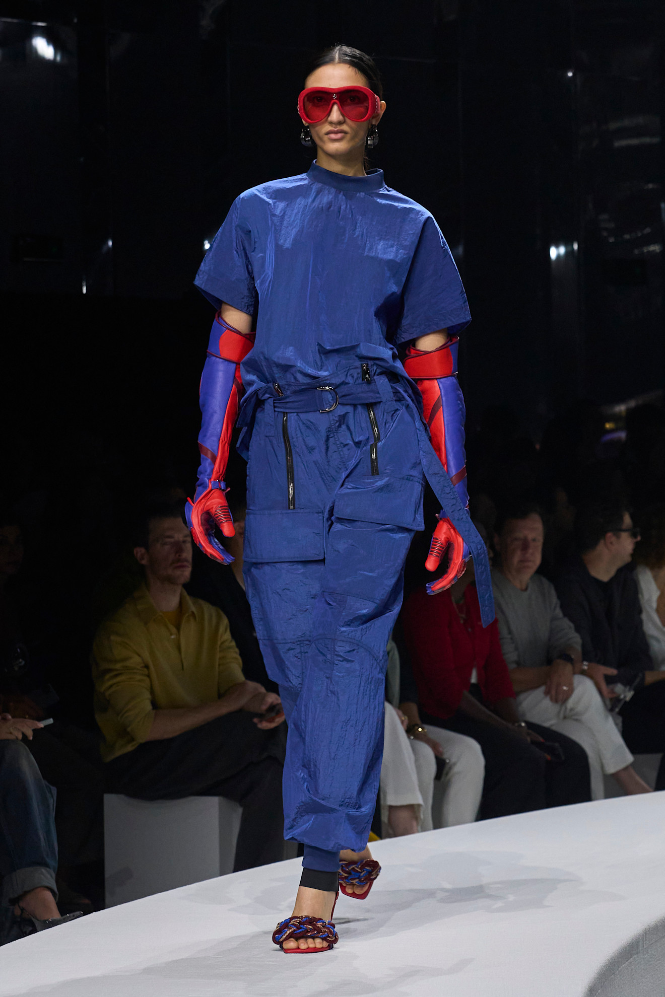 Модель в ярком синем комбинезоне с объемными карманами, дополненном красными перчатками и крупными очками, создает футуристичный образ.