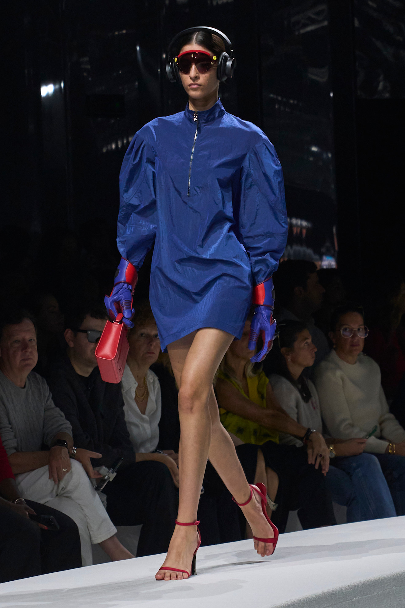 Модель в ярком синем платье с объемными рукавами, красных сандалиях и стильных наушниках, держит мини-сумку в тон.