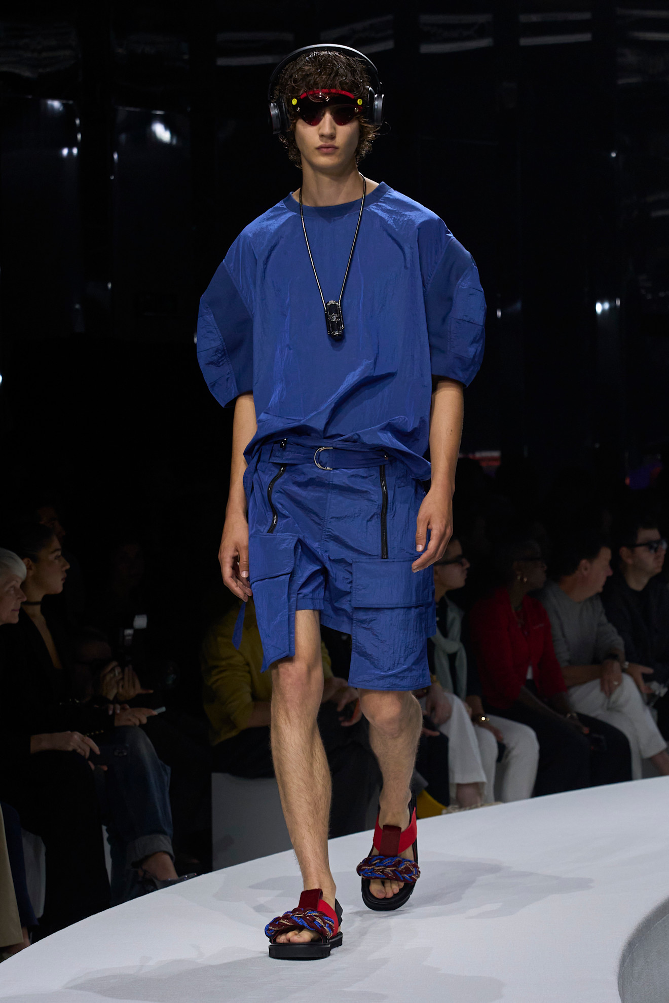 Модель в ярком синем комплекте с объемной футболкой и шортами, дополненными функциональными карманами, завершает образ яркими сандалиями.