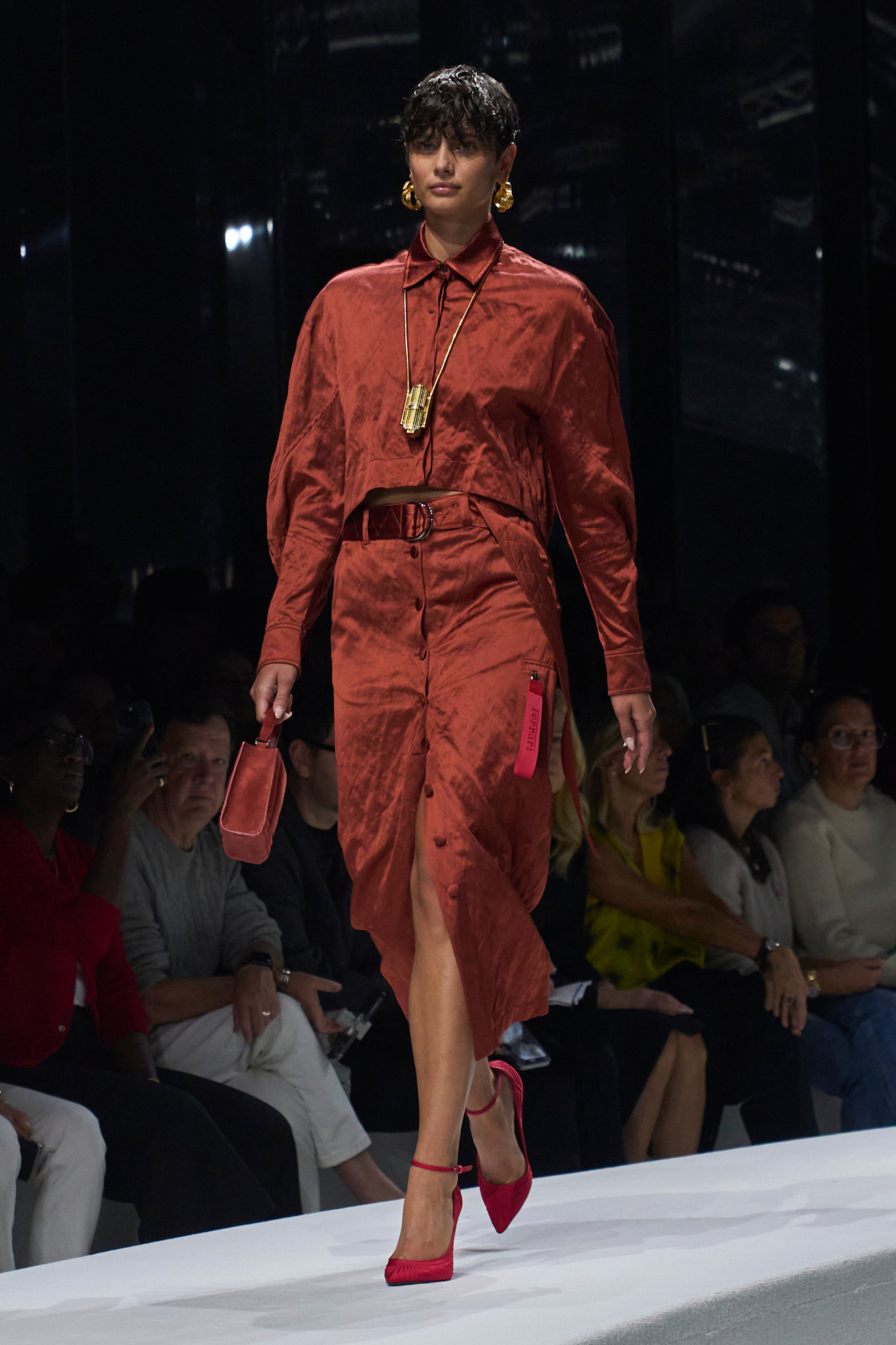 Модель в атласном платье терракотового оттенка с асимметричным разрезом, дополненном яркими аксессуарами и элегантными туфлями на каблуке...