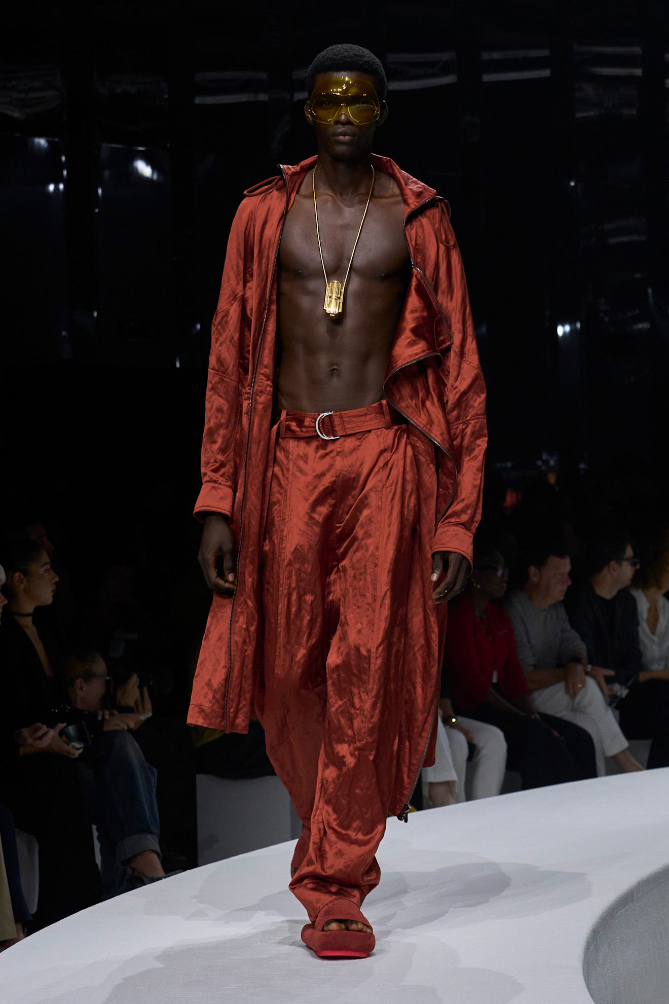 Модель в ярком оранжевом комплекте из блестящей ткани, дополненном крупными золотыми аксессуарами и солнцезащитными очками, уверенно шагает по подиуму....