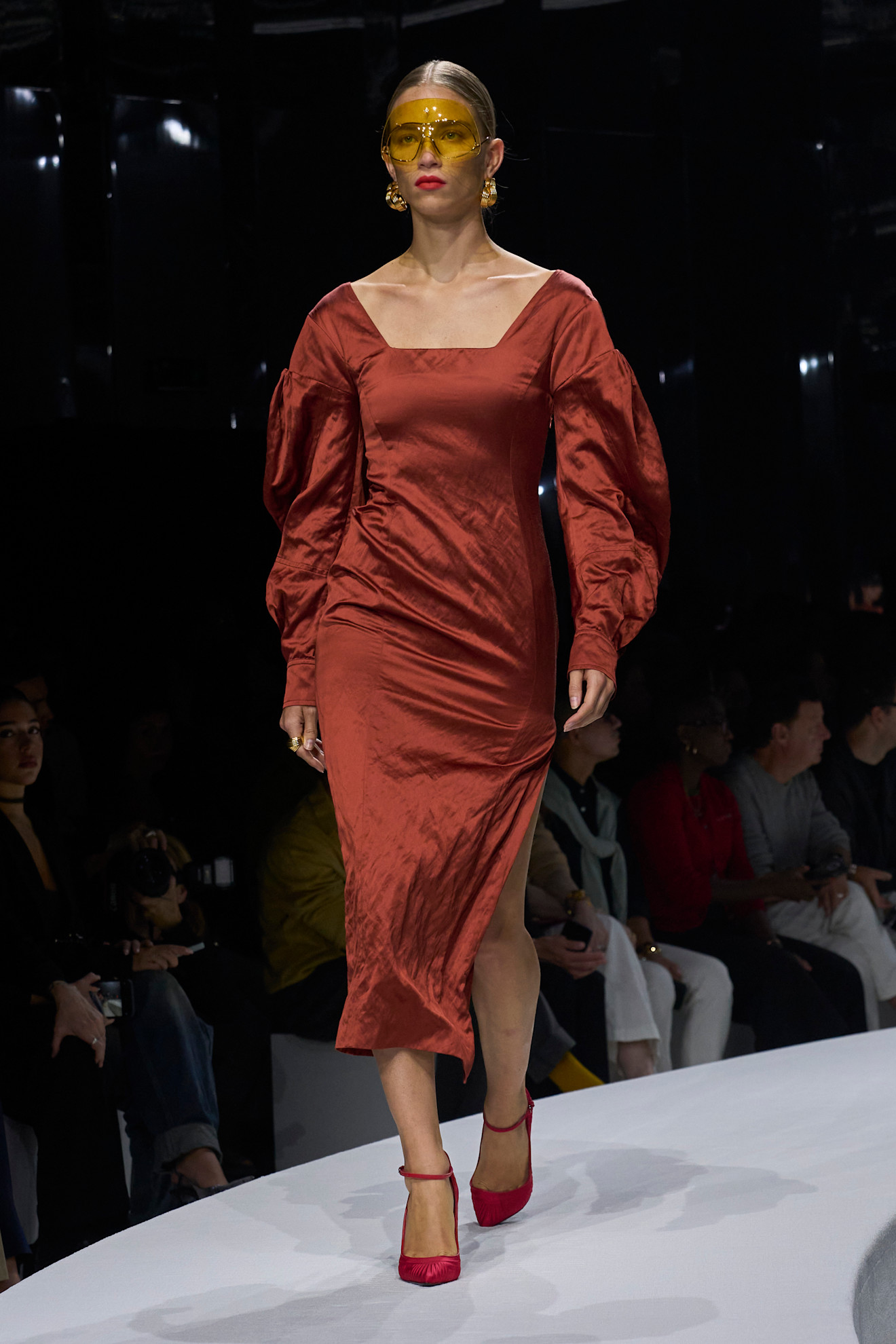 Модель в атласном терракотовом платье с объемными рукавами, ярких желтых очках и розовых туфлях, уверенно шагает по подиуму.