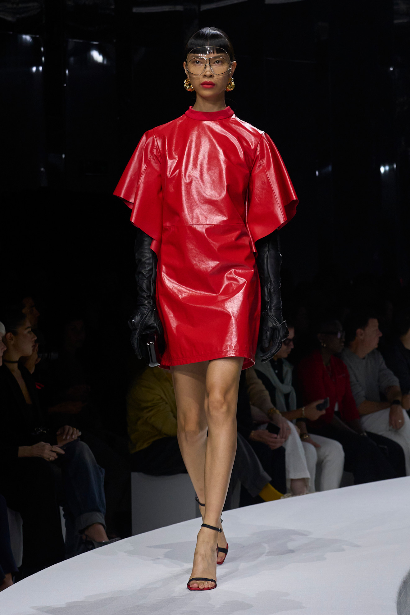 Яркое красное платье из лакированной кожи с объемными рукавами, дополненное черными перчатками и крупными золотыми серьгами, создает эффектный образ для весенне-летнего сезона 2024 от Ferrari...