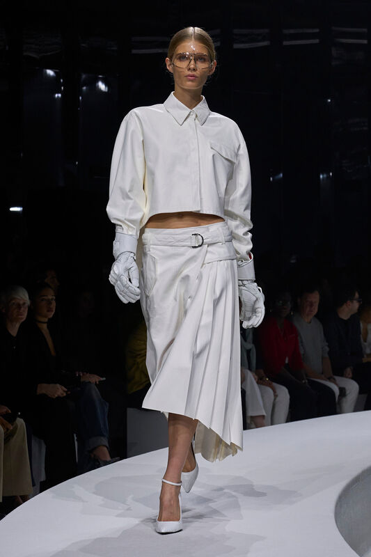 Модель в белоснежном комплекте с укороченной рубашкой и асимметричной юбкой, дополненная перчатками и стильными очками, создает атмосферу элегантной современности....