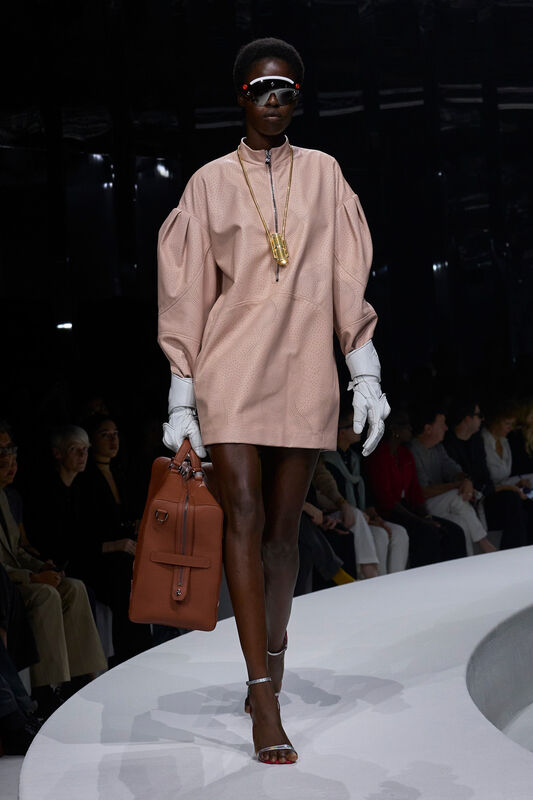 Модель в розовом платье с объемными рукавами, дополненном белыми перчатками и стильными очками, несет коричневую сумку.