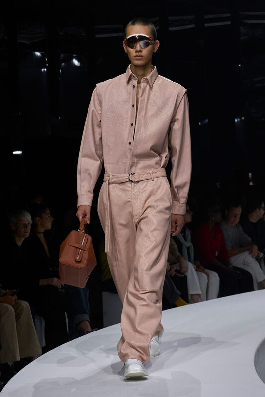 Мужская коллекция Ferrari весна/лето 2024: нежно-розовая рубашка с перфорацией и свободные брюки, дополненные стильной сумкой и очками.