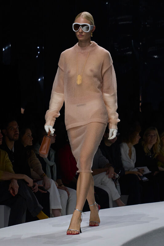 Модель в полупрозрачном розовом наряде с длинными рукавами и облегающей юбкой, дополненная крупными очками и стильной сумочкой.