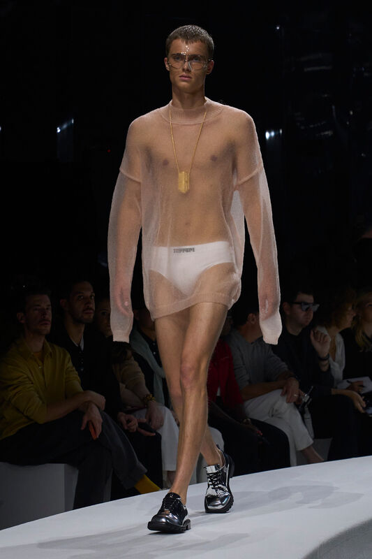 Модель в полупрозрачном розовом свитере и белых плавках демонстрирует смелый стиль с металлическими аксессуарами на фоне зрителей.