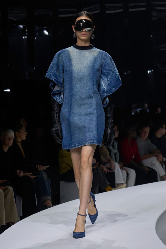 Девушка в асимметричном джинсовом платье с объемными рукавами, черных кожаных перчатках и стильных очках, уверенно шагает по подиуму.