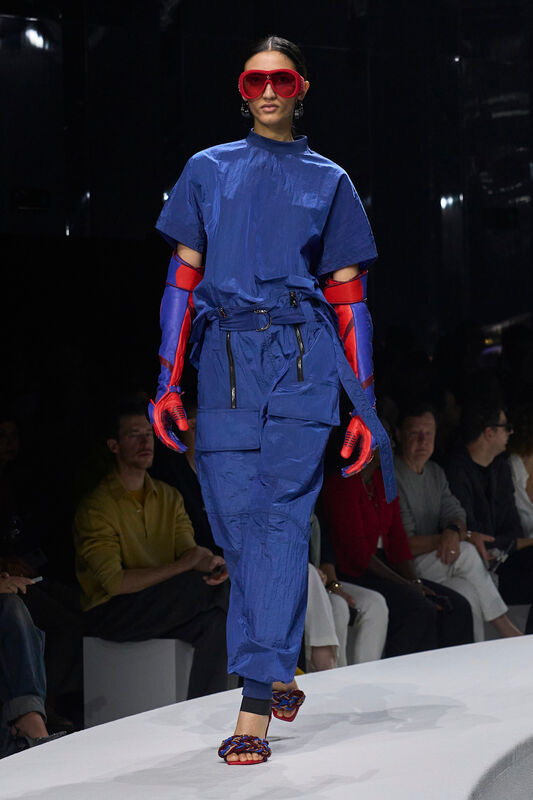 Модель в ярком синем комбинезоне с объемными карманами, дополненном красными перчатками и крупными очками, создает футуристичный образ.