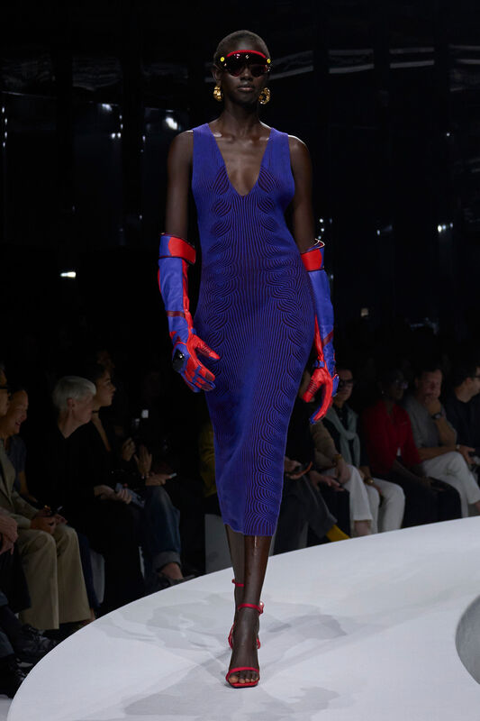 Модель в облегающем синем платье с волнистым узором, дополненном яркими красными перчатками и обувью на высоком каблуке, уверенно шагает по подиуму....
