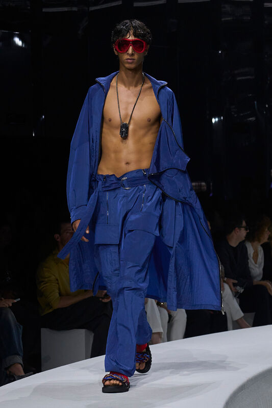 Модель в ярком синем комплекте с открытой грудью и объемным плащом, дополненным крупными красными очками и стильными сандалиями.